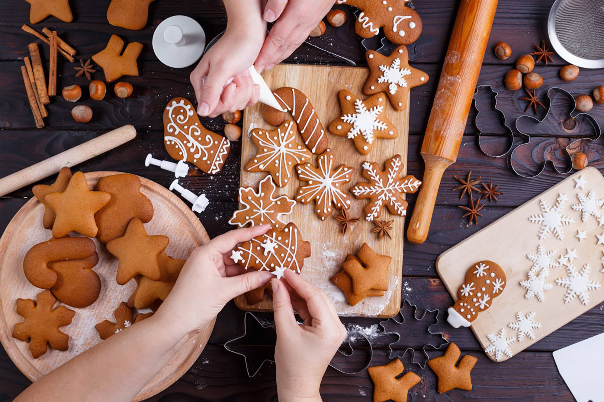 2 Personen verzieren Lebkuchenplätzchen mit weißem Zuckerguss und Fondantsternen auf Holzbrett.