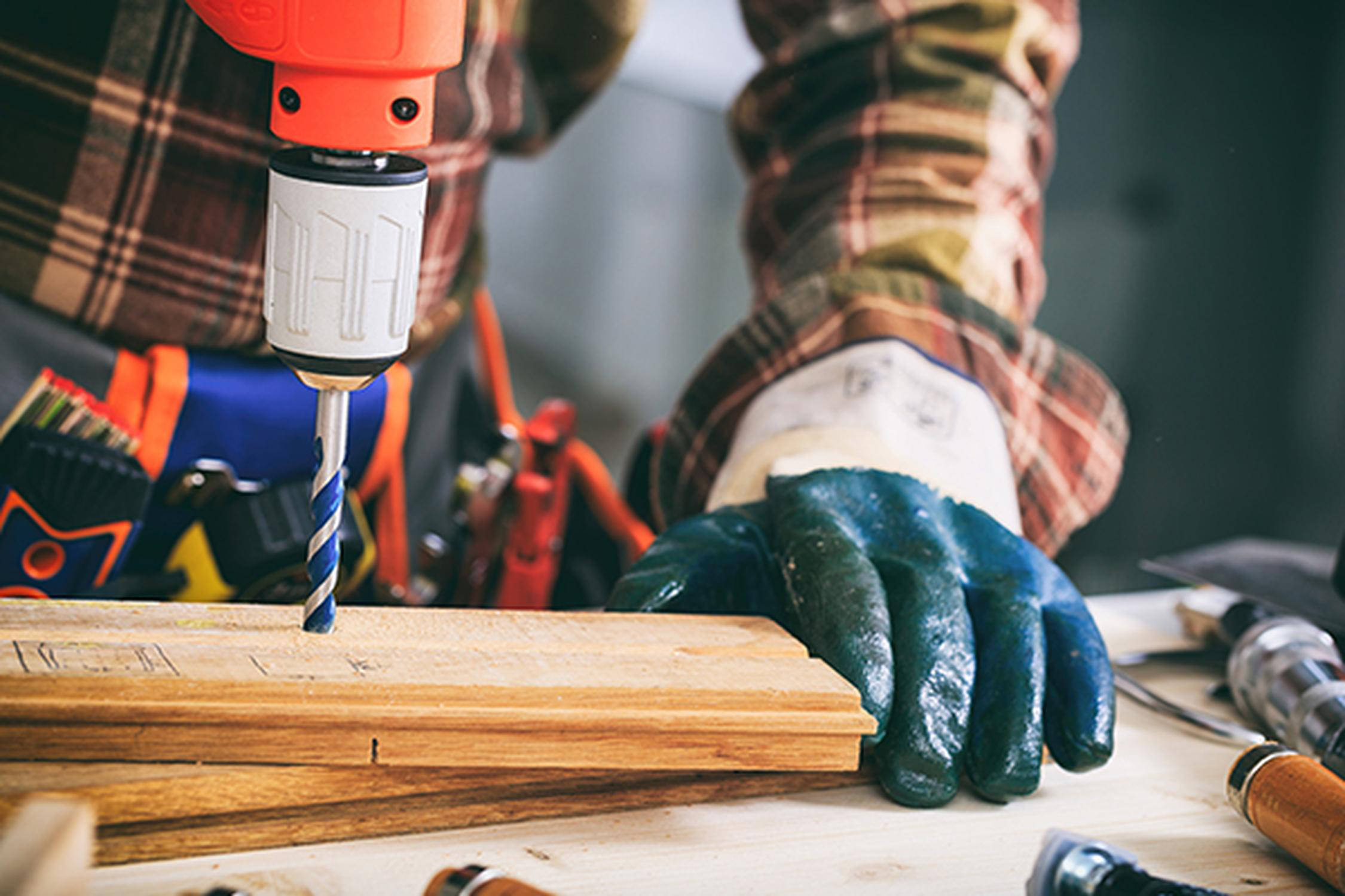 Bohrmaschine bohrt ein Loch in ein Holzbrett, das von einer Hand mit Handschuh gehalten wird