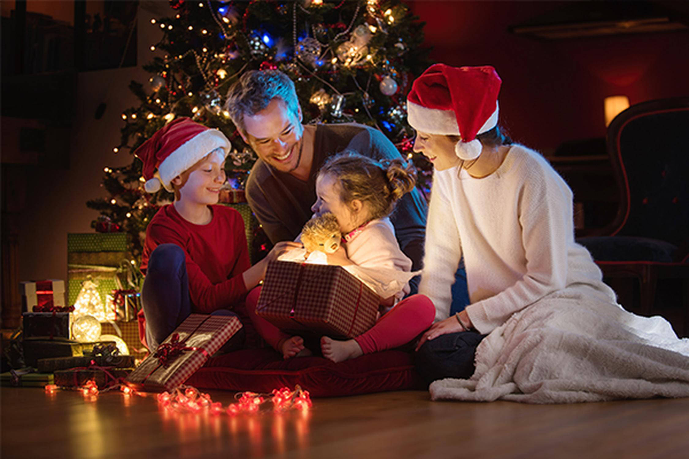 ein Mann, eine Frau mit roter Weihnachtsmütze und zwei Kinder mit je einer Weihnachtsmütze vor beleuchteten Christbaum