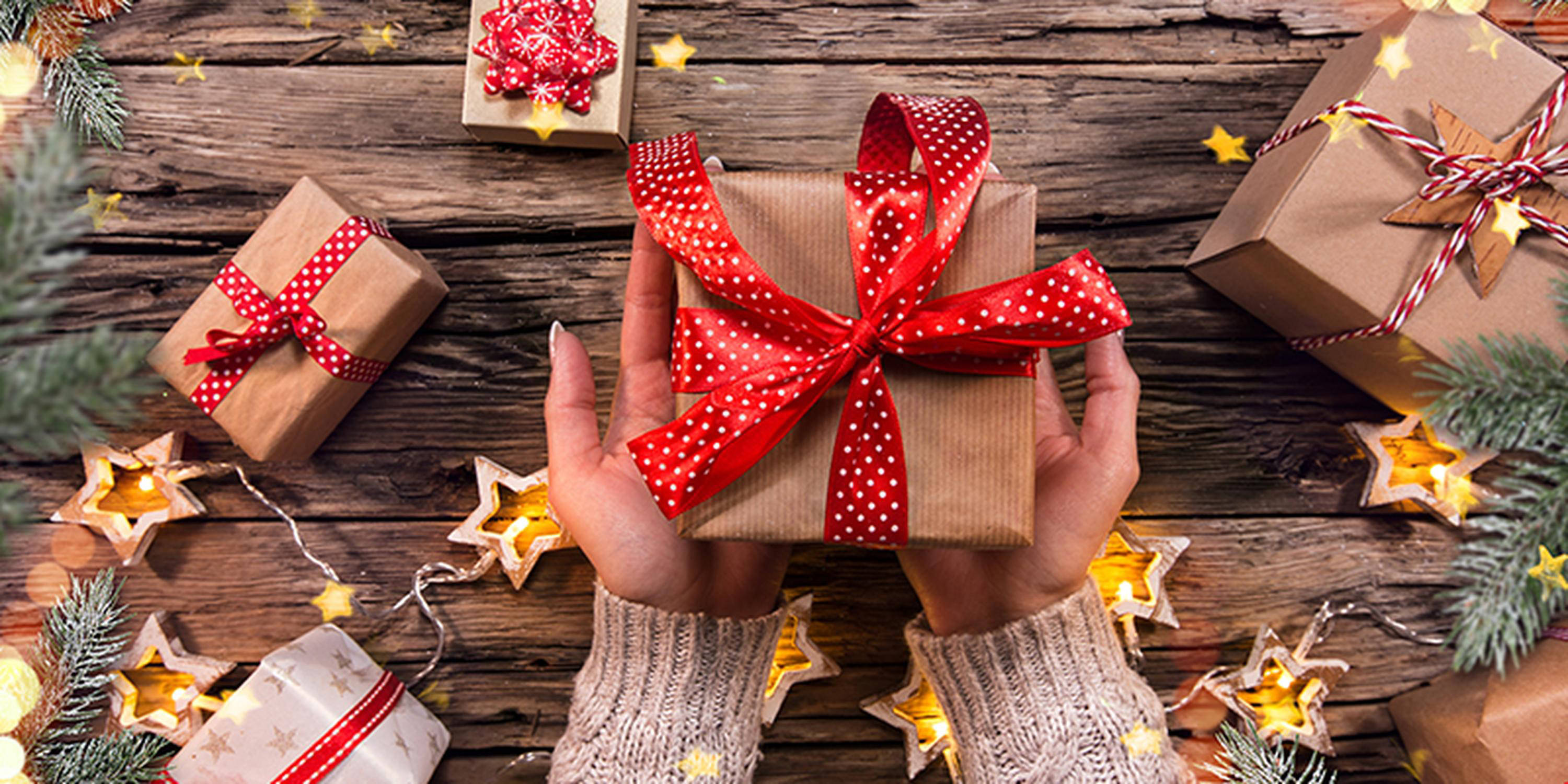 Frauenhände halten ein in braunem Papier eingewickeltes Geschenk mit rot-weißer Schleife. Im Hintergrund sind weitere Ge