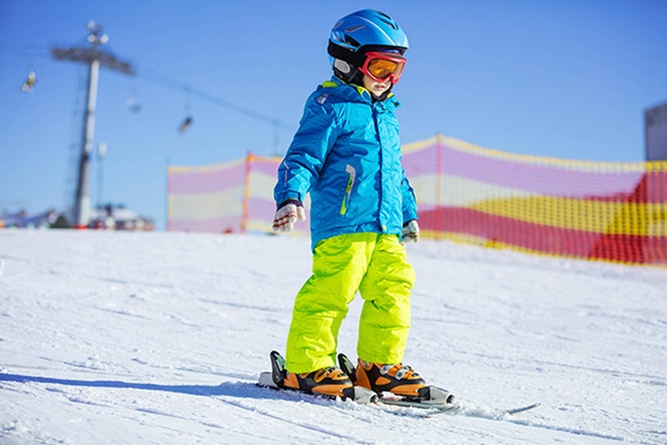 Kinde mit blauem Helm, blauer Skijacke und grüner Skihose fährt auf Skiern