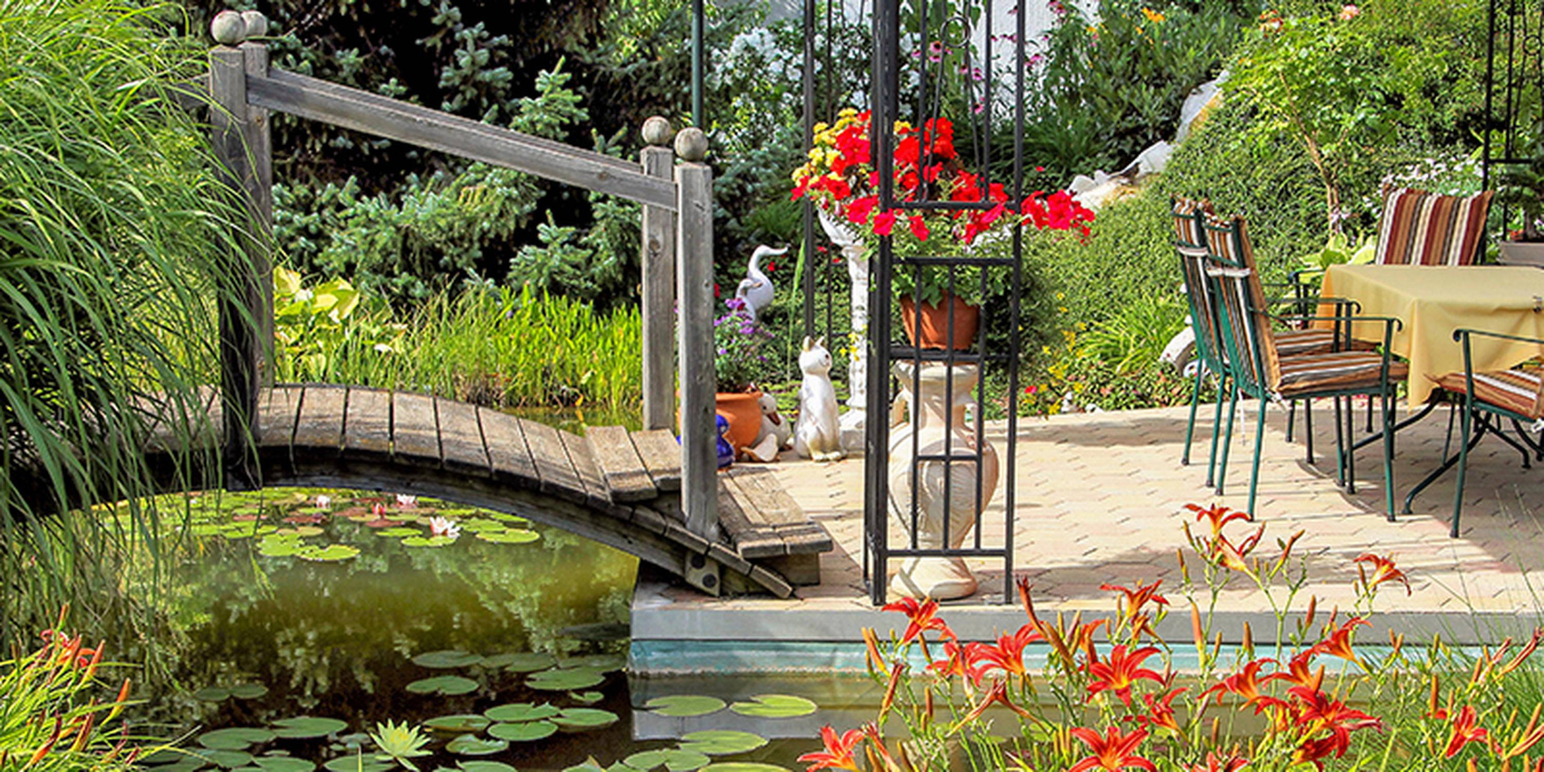 Gartenstühle und ein Tisch stehen auf einer Garteninsel, die über eine kleine Bogenbrücke über Wasser erreichbar ist.