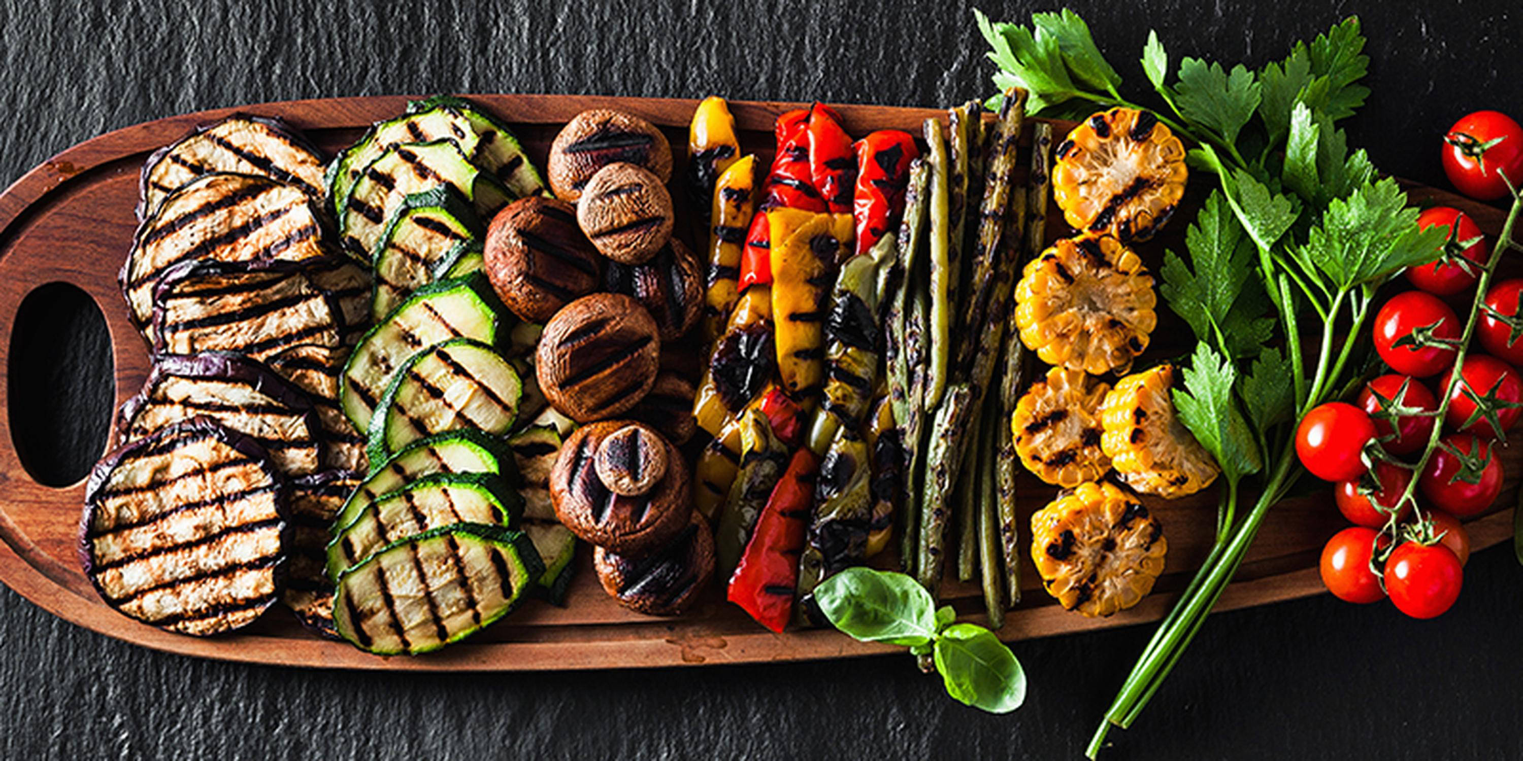 Auf einem großen Holzbrett liegen gegrillte Melanzani, Zucchini, Pilze, Paprika, Bohnen, Maiskolben und Rispentomaten. Daneben stehen ein Salz- und Pfefferstreuer.