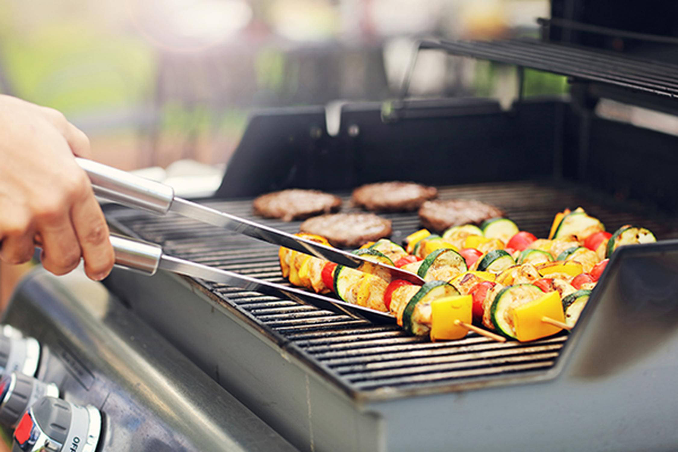Auf dem Bild ist zu sehen, wie jemand einen Grillspieß auf einem Gasgrill wendet. Auf dem Gasgrill sind zudem Fleischpat