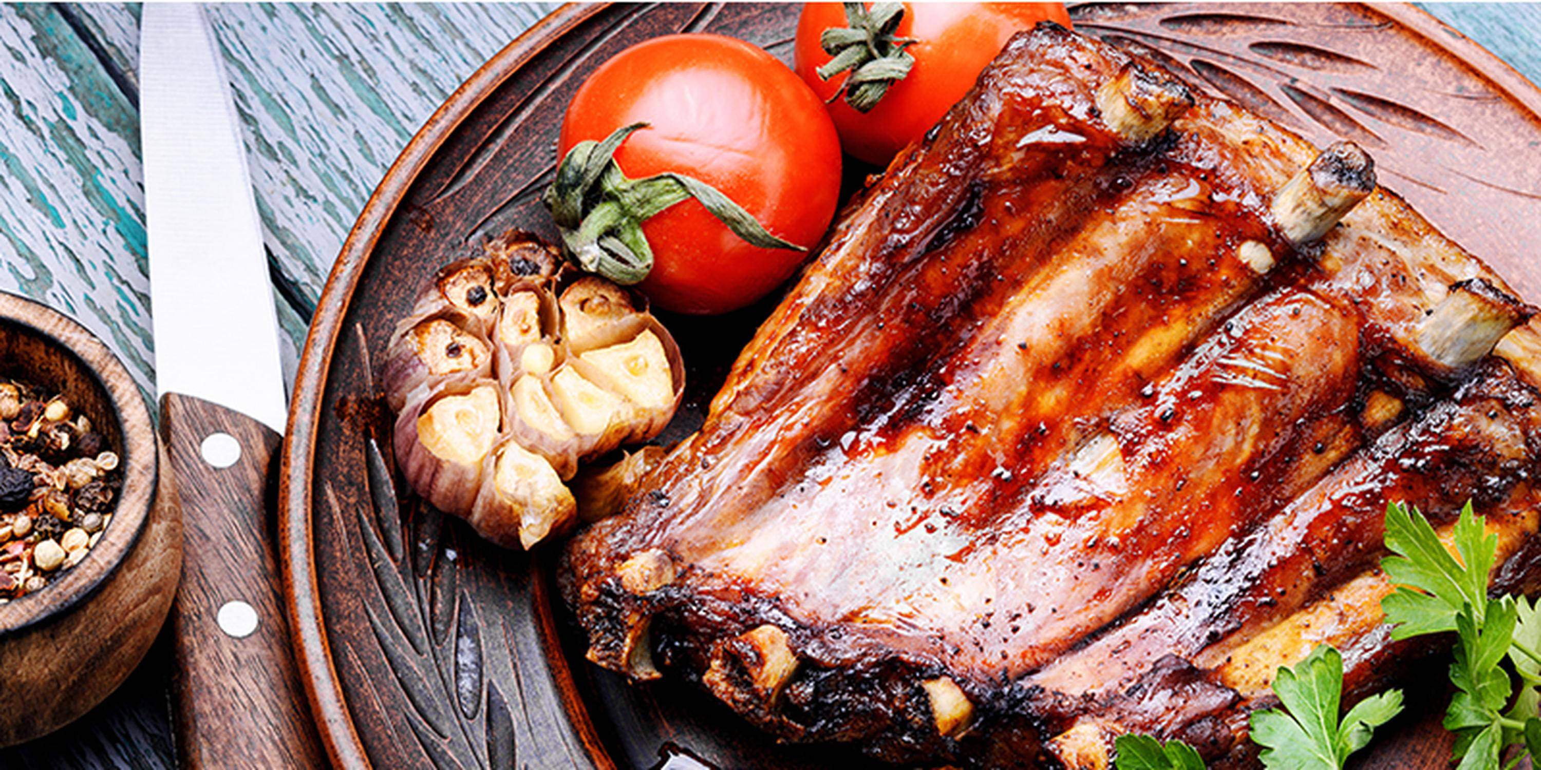 Spareribs mit Knoblauch auf einem Holzteller. Links daneben liegt ein Messer und eine Holzschale mit Gewürzen.
