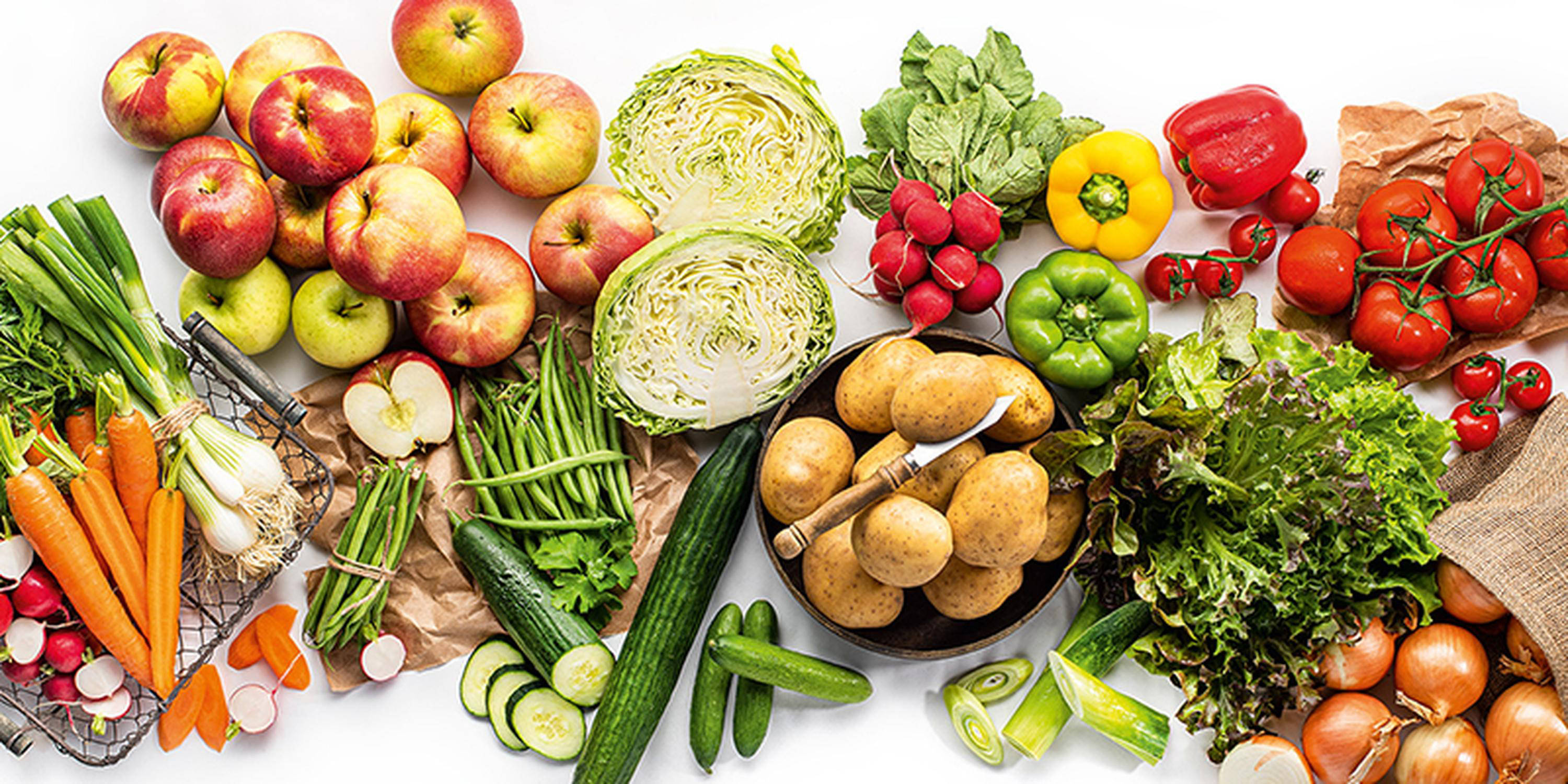 Auf weißem Hintergrund liegt unterschiedliches Obst und Gemüse. In der Mitte ist eine Schale mit Kartoffeln. 