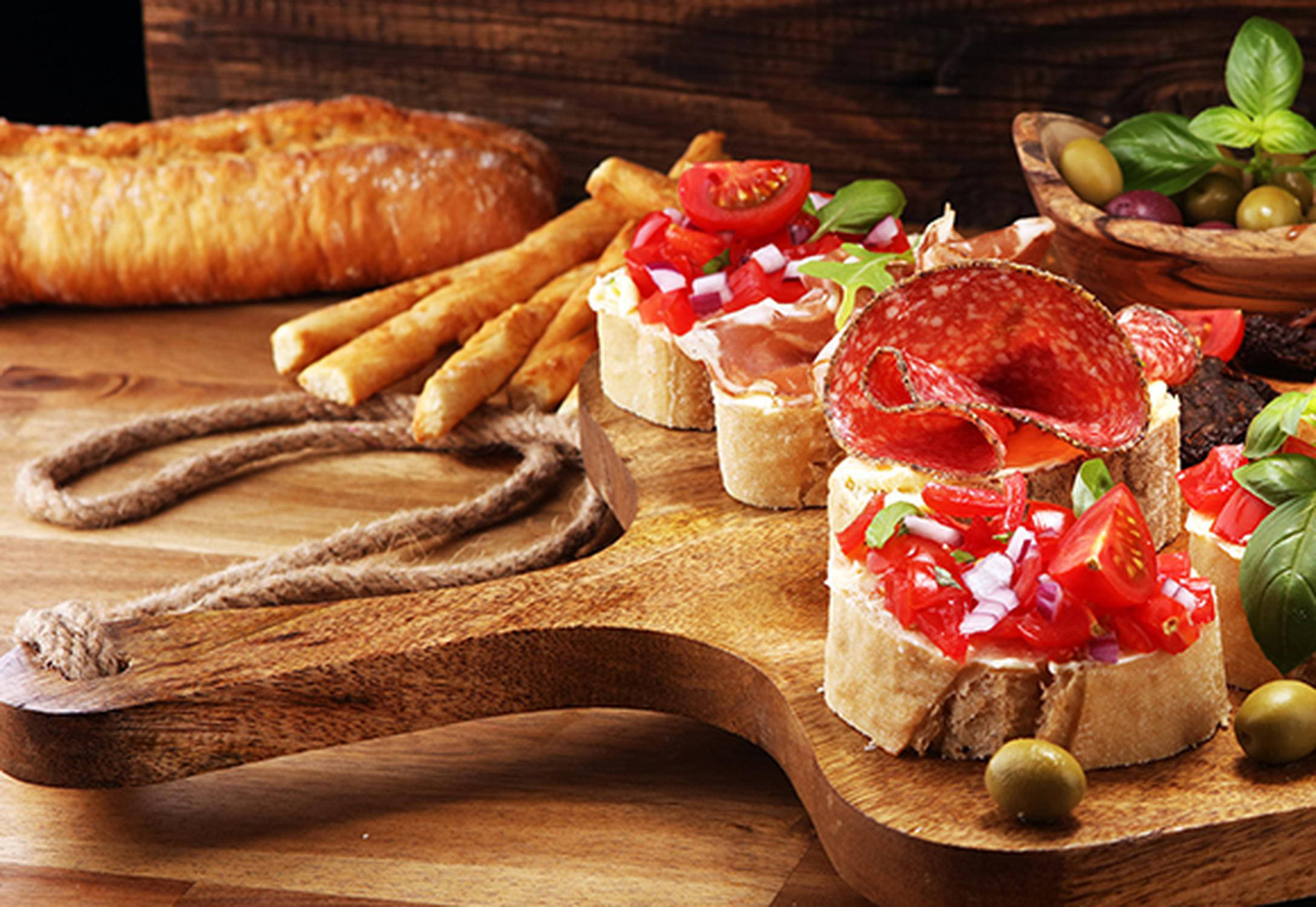 Ein Jausenbrett aus Holz auf dem mehrere mit Tomaten und Salami belegte Brötchen liegen. 