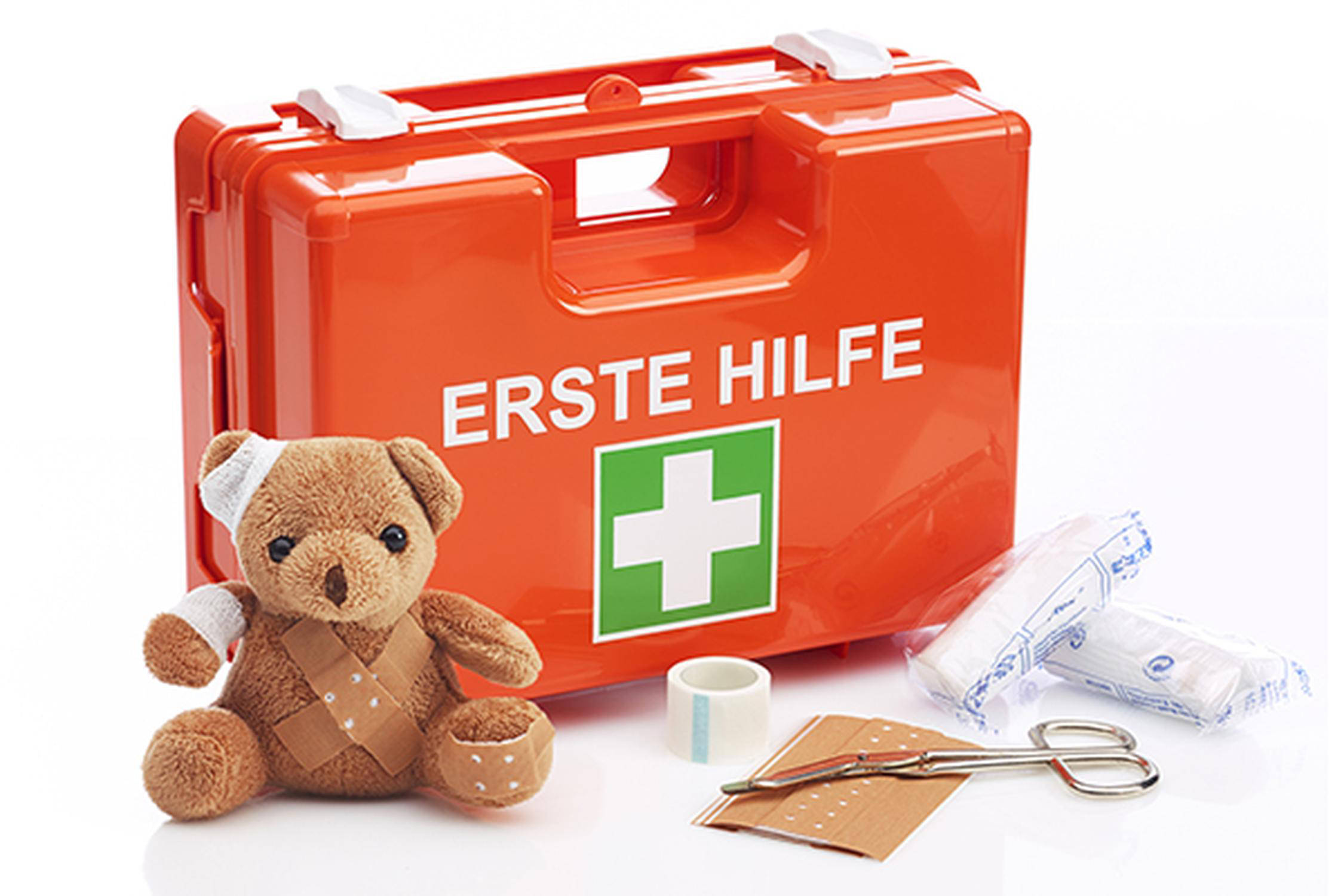 Ein oranger Erste Hilfe Koffer. Davor sitzt ein bandagierter und mit Pflastern beklebter Teddybär. Rechts daneben eine Verbandsschere, zwei Verbände und ein Streifen Pflaster.
