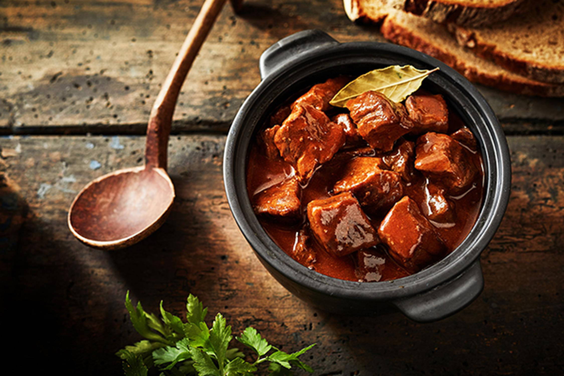 auf einem Holzuntergrund steht ein Topf mit Gulasch und ein Löffel und Brotscheiben daneben