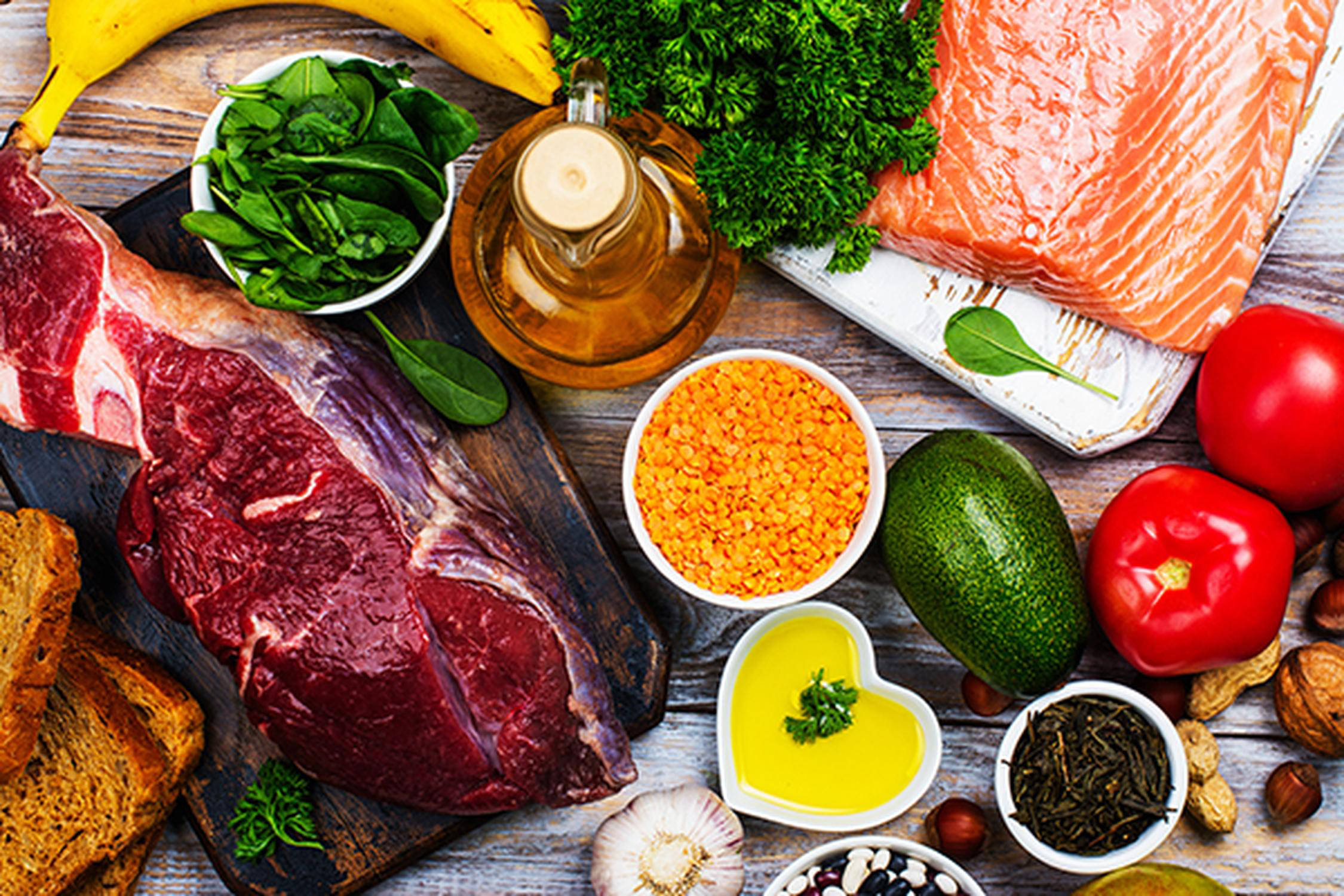 Holzunterlage mit Fleisch, Brot, Fisch, Kräuter, Gemüse, Banane und Öl