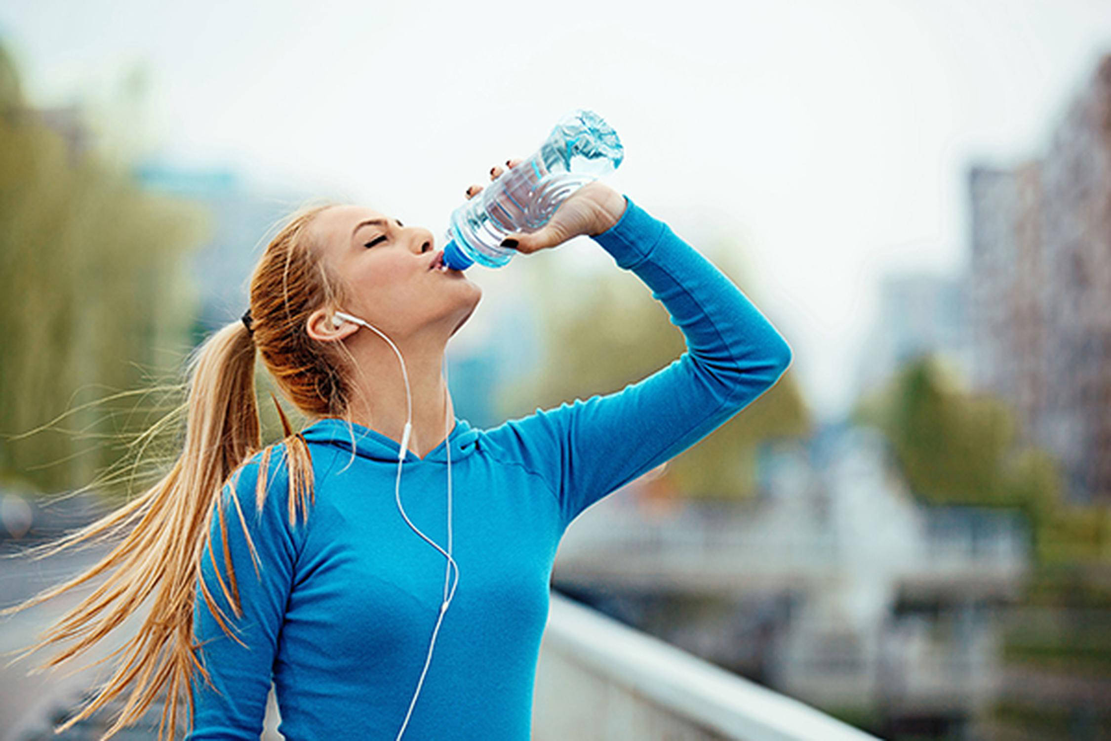 Frau mit Zopf, blauem Shirt und Kopfhörern trinkt bei einer Wasserflasche