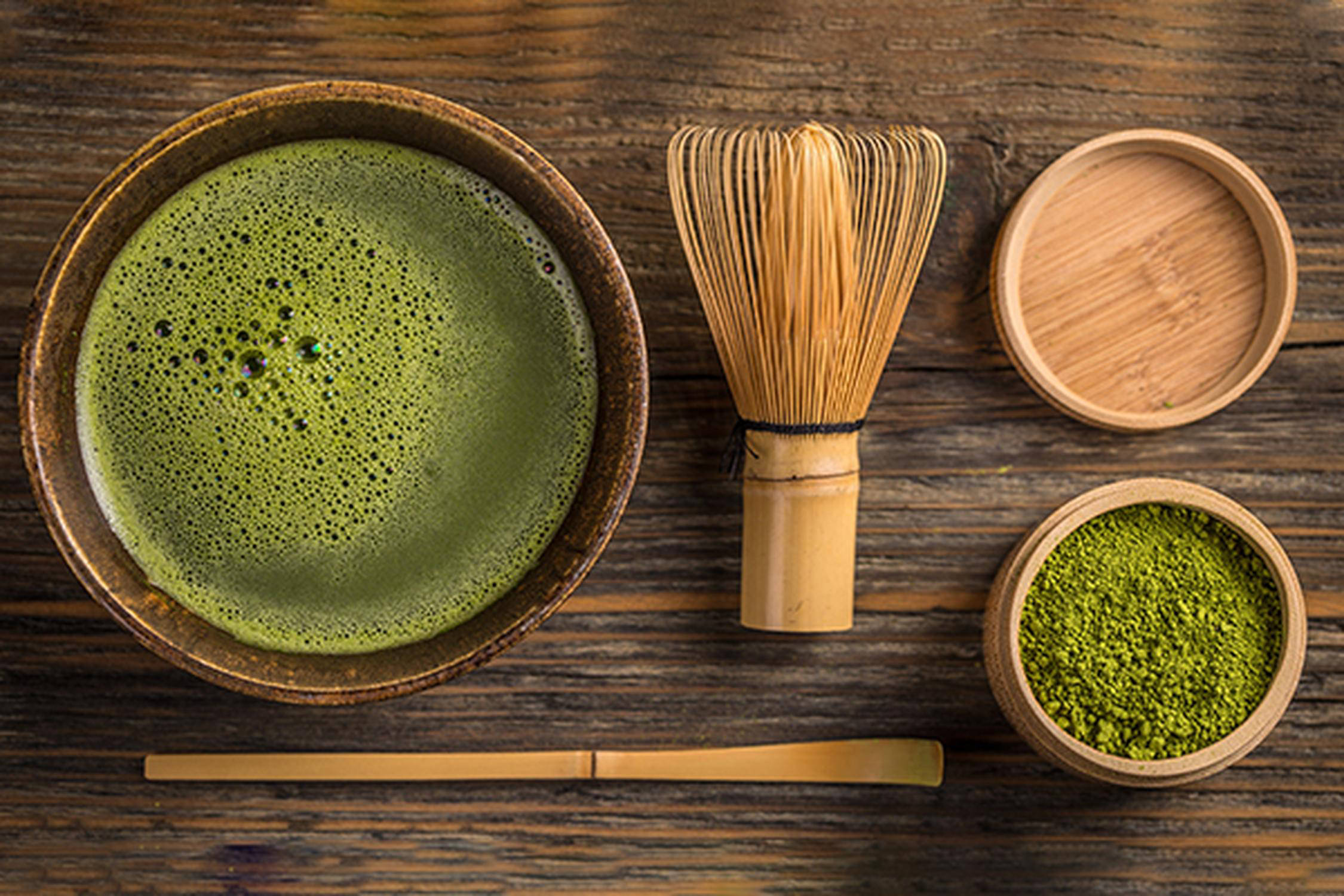 Matcha in Flüssigkeit aufgelöst in einer Schüssel und als Pulver in einer anderen Schüssel, daneben ein Deckel, Stäbchen