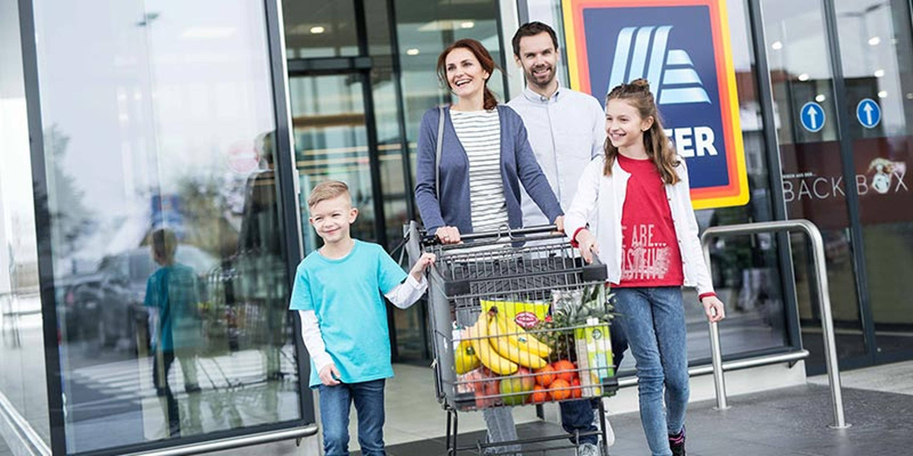 Vierköpfige Familie verlässt glücklich mit Einkaufswagen ein HOFER Geschäft.
