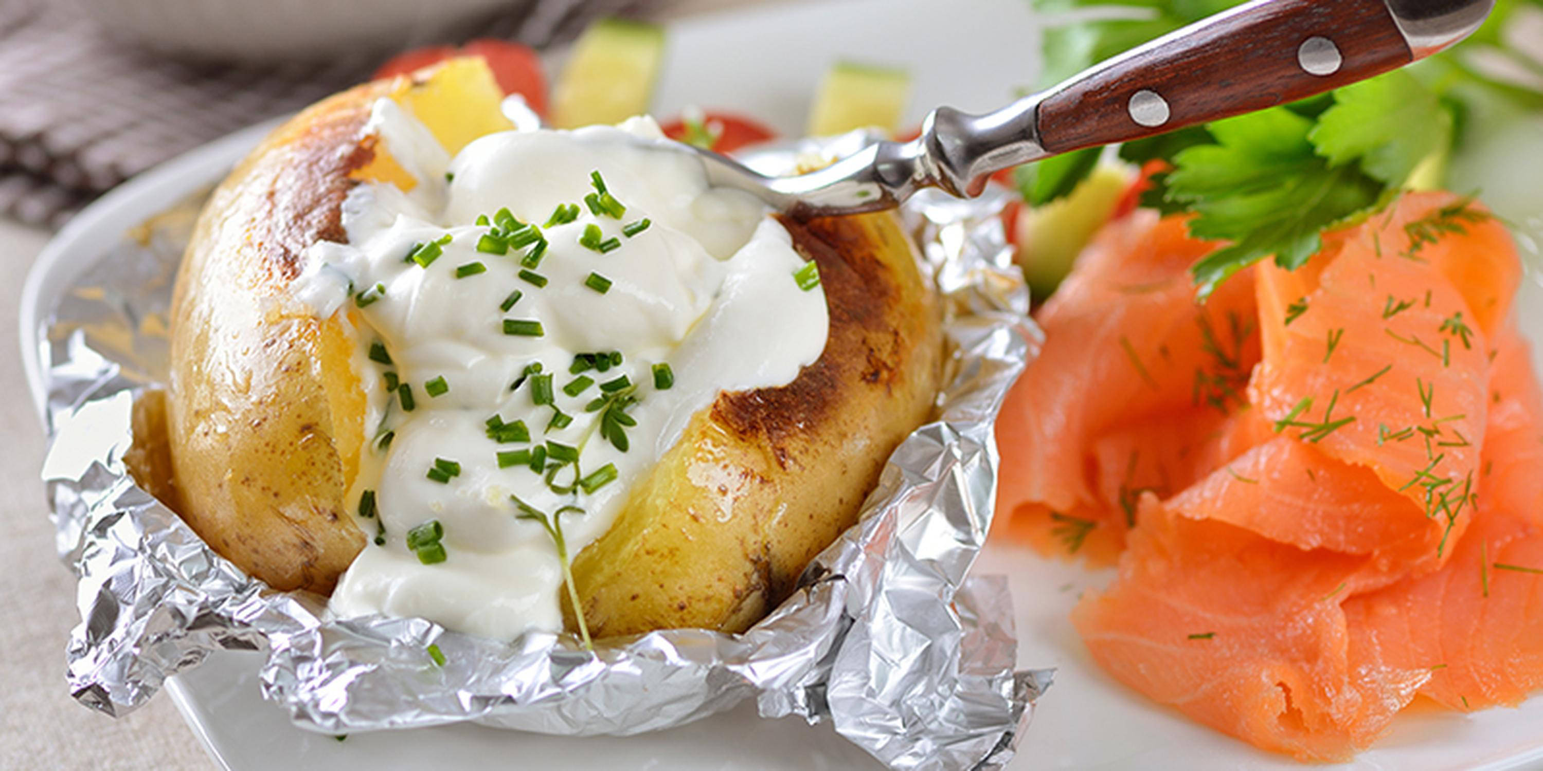 Eine Ofenkartoffel mit Soße und Schnittlauch auf Aluminiumfolie. Daneben etwas Räucherlachs.