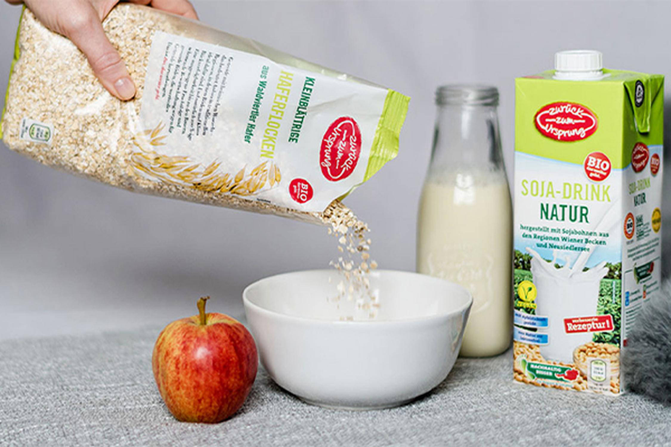 Aus einer Packung werden Haferflocken in eine weiße Schüssel geschüttet, daneben liegen ein Apfel und Milch