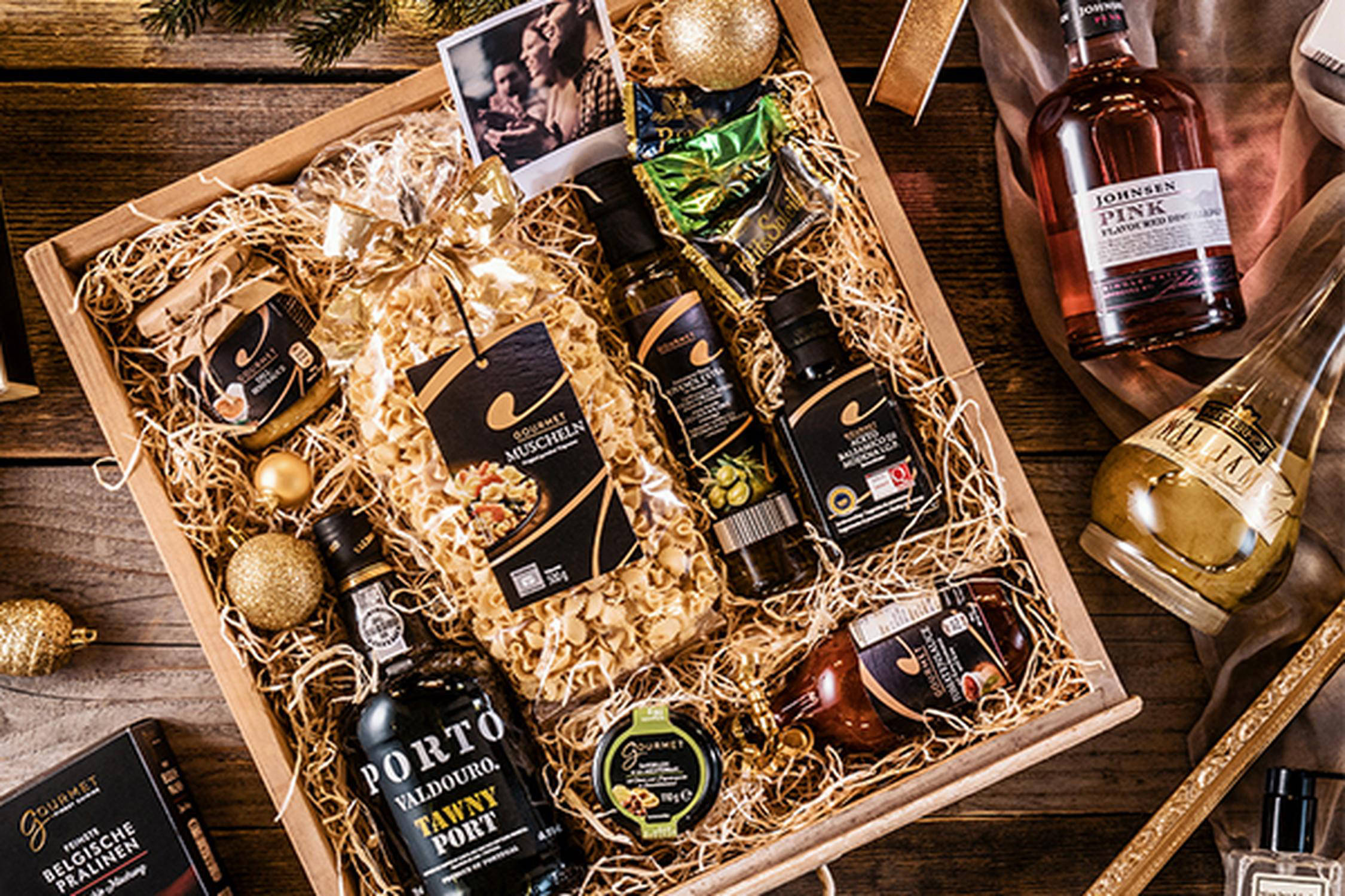 Geschenkbox mit verschiedenen Flaschen, goldenen Christbaumkugeln, einer Packung Nudeln, daneben liegen zwei Flaschen