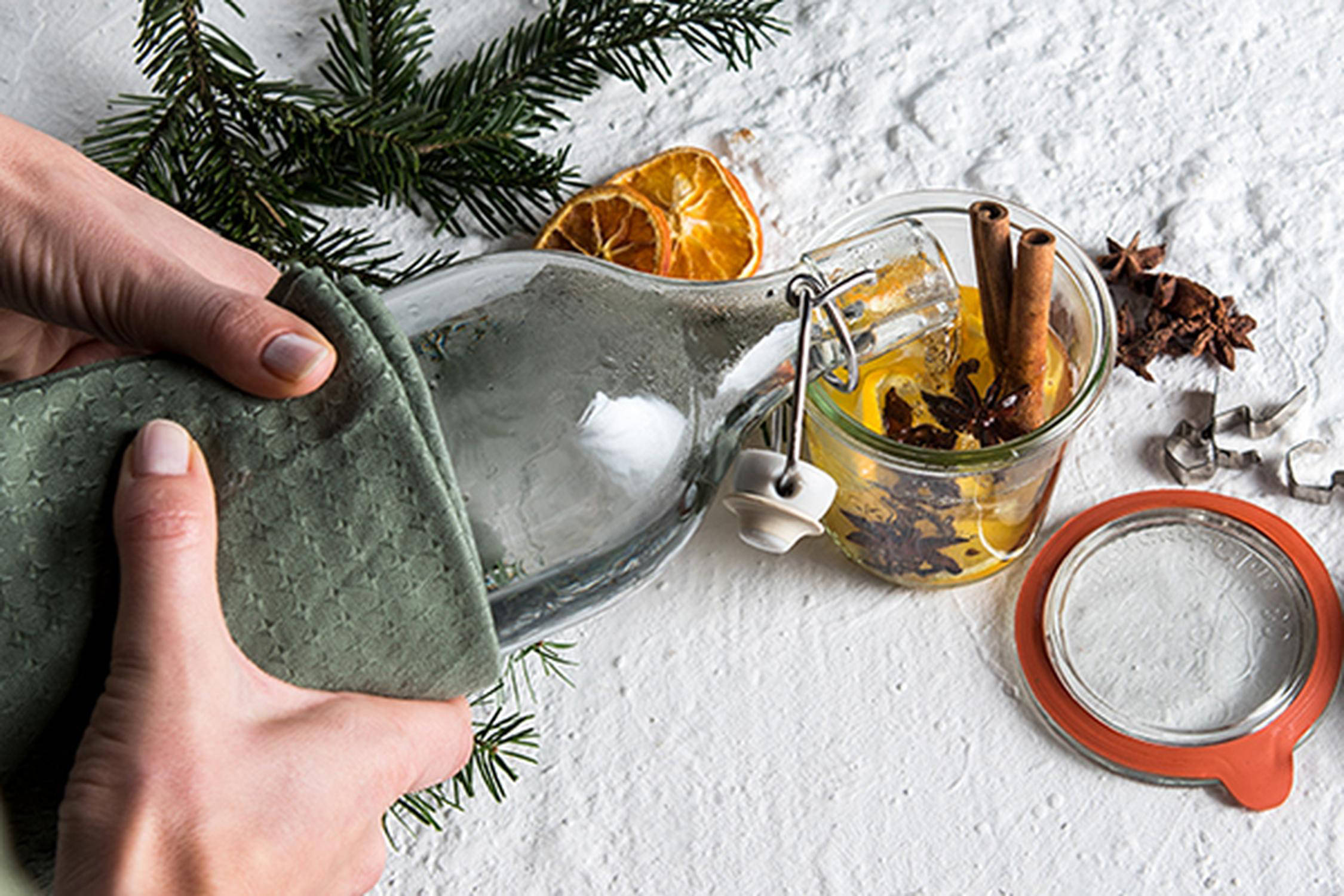 zwei Hände halten mit einem grünen Tuch eine Glasflasche und leeren den Inhalt in ein Vorratsglas mit Zimt, Nelken und O
