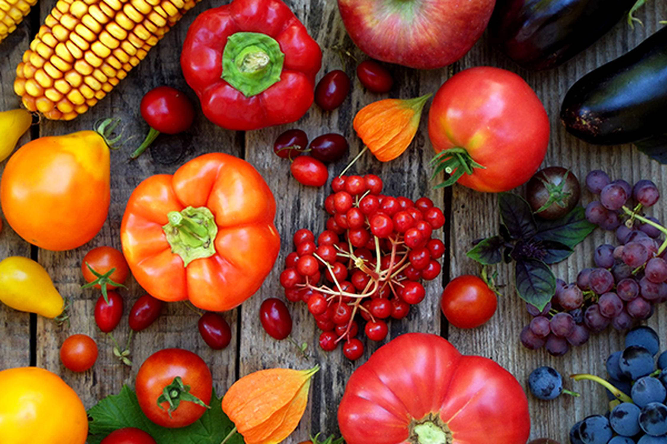 Mais, Paprika, Äpfel, Tomaten und Beeren liegen auf einem Holzuntergrund.