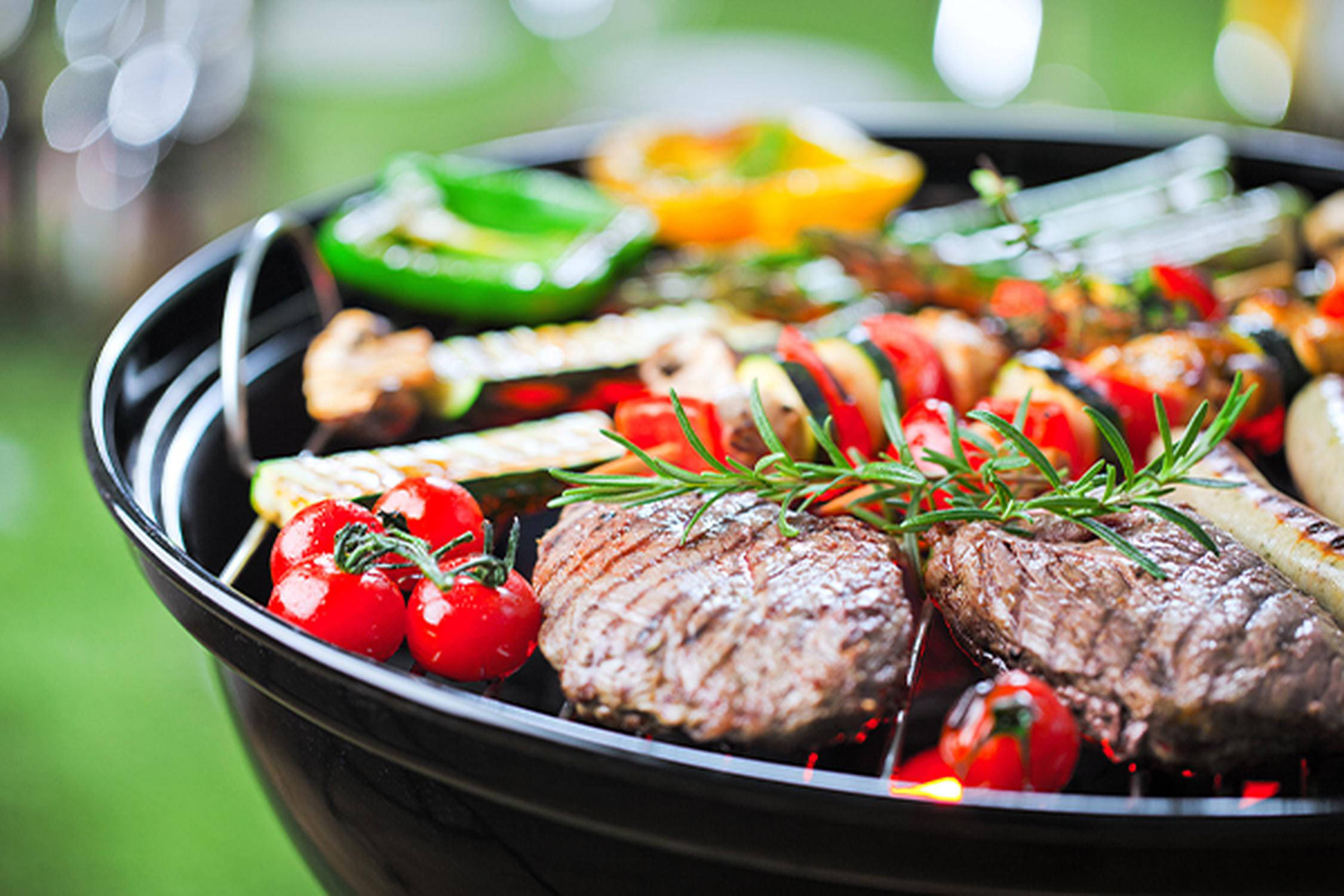 Auf dem Bild ist ein Kugelgrill mit diversen Grillgut ersichtlich. Auf dem Grill liegen Tomaten, Fleisch, Rosmarin, Papr