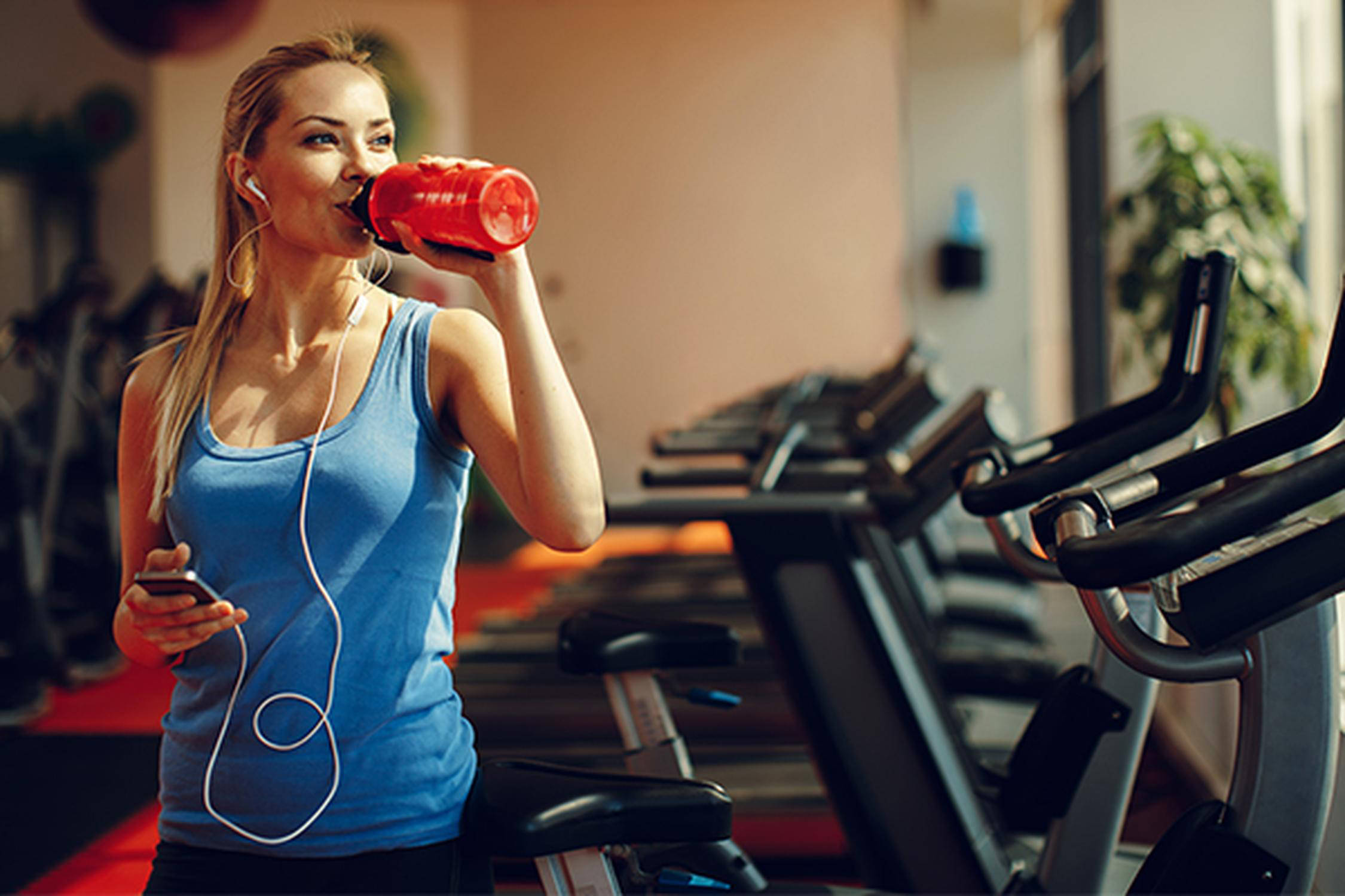Eine Frau trinkt von einer roten Trinkflasche und steht neben einem Heimtrainer