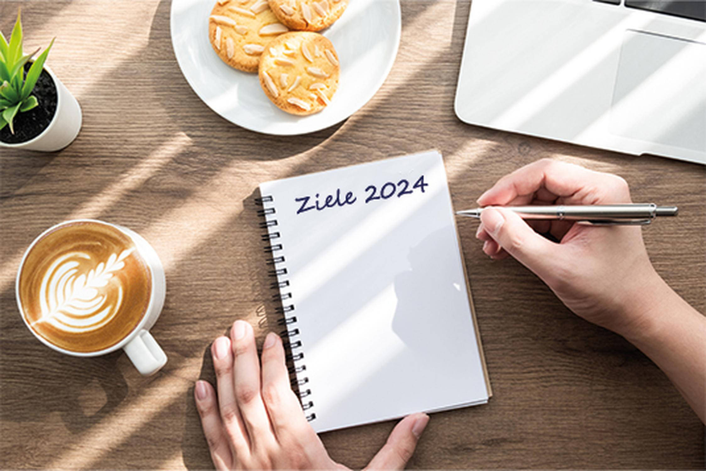 weißes Blatt Papier mit den Worten "Ziele 2024) eine Hält eine Stift, daneben eine Tasse mit Kaffe, ein Teller mit Kekse