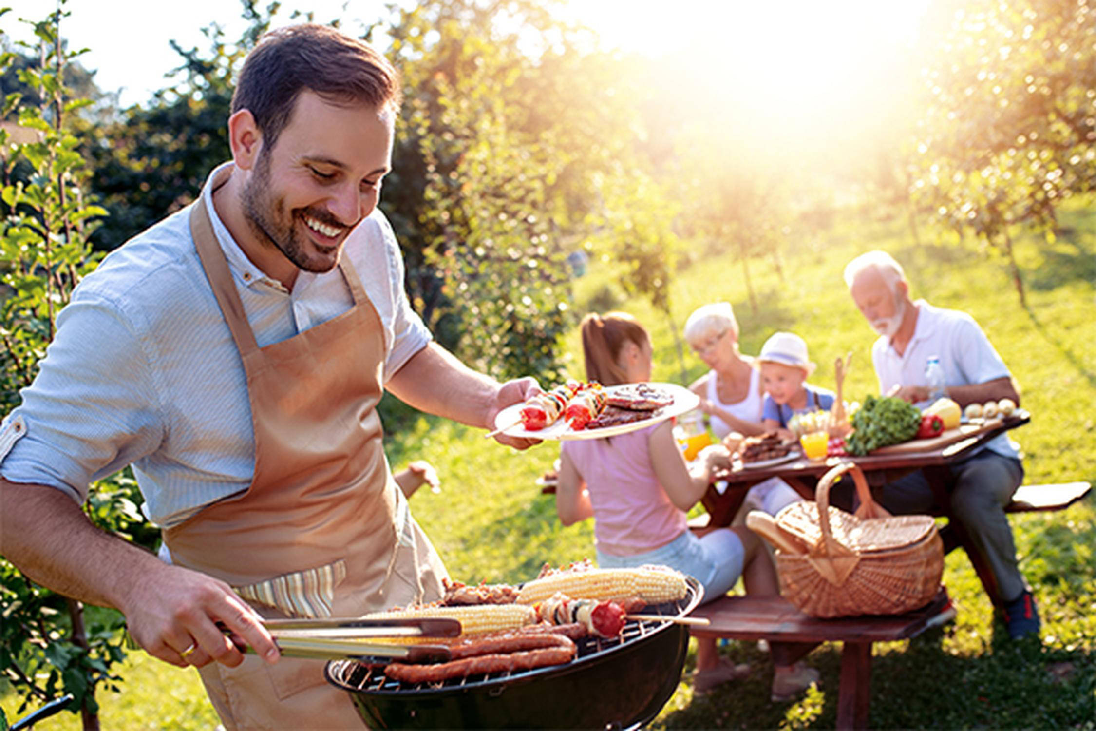 Mann steht am Griller und grillt Würstchen und Mais, im Hintergrund sitzen drei Erwachsene und ein Kind an einem Tisch