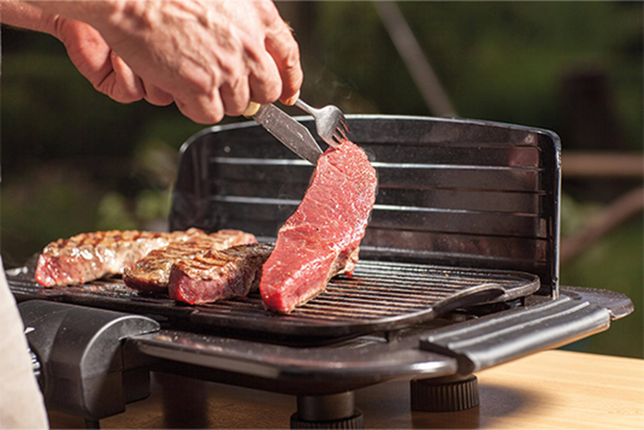 Auf dem Bild ist zu sehen, wie jemand ein Steak auf einem Elektrogrill wendet. Insgesamt sind vier Steaks auf dem Grill.
