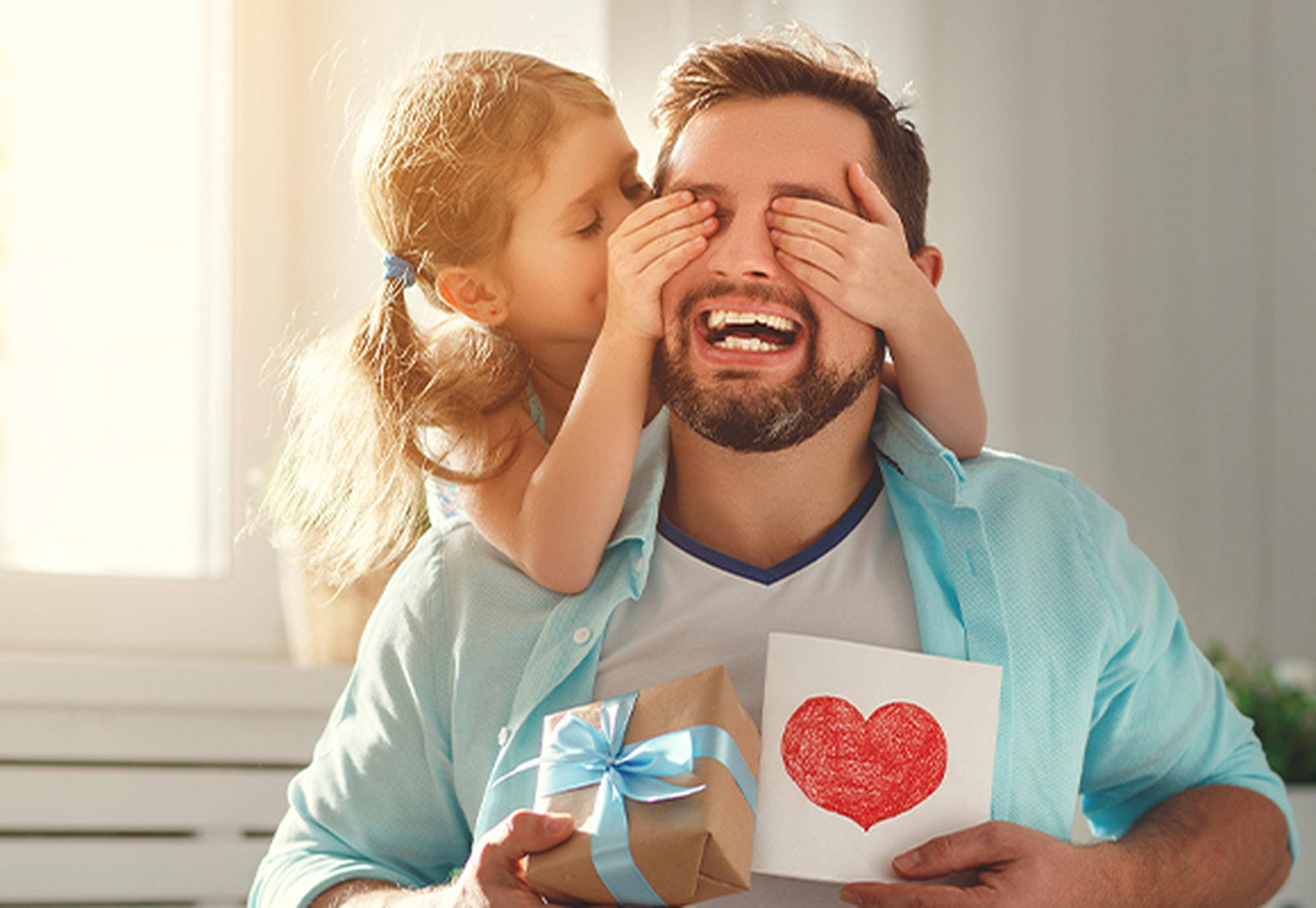 Mann hält eine Karte und ein kleines Päckchen in den Händen und ein Mädchen hält ihm mit beiden Händen die Augen zu