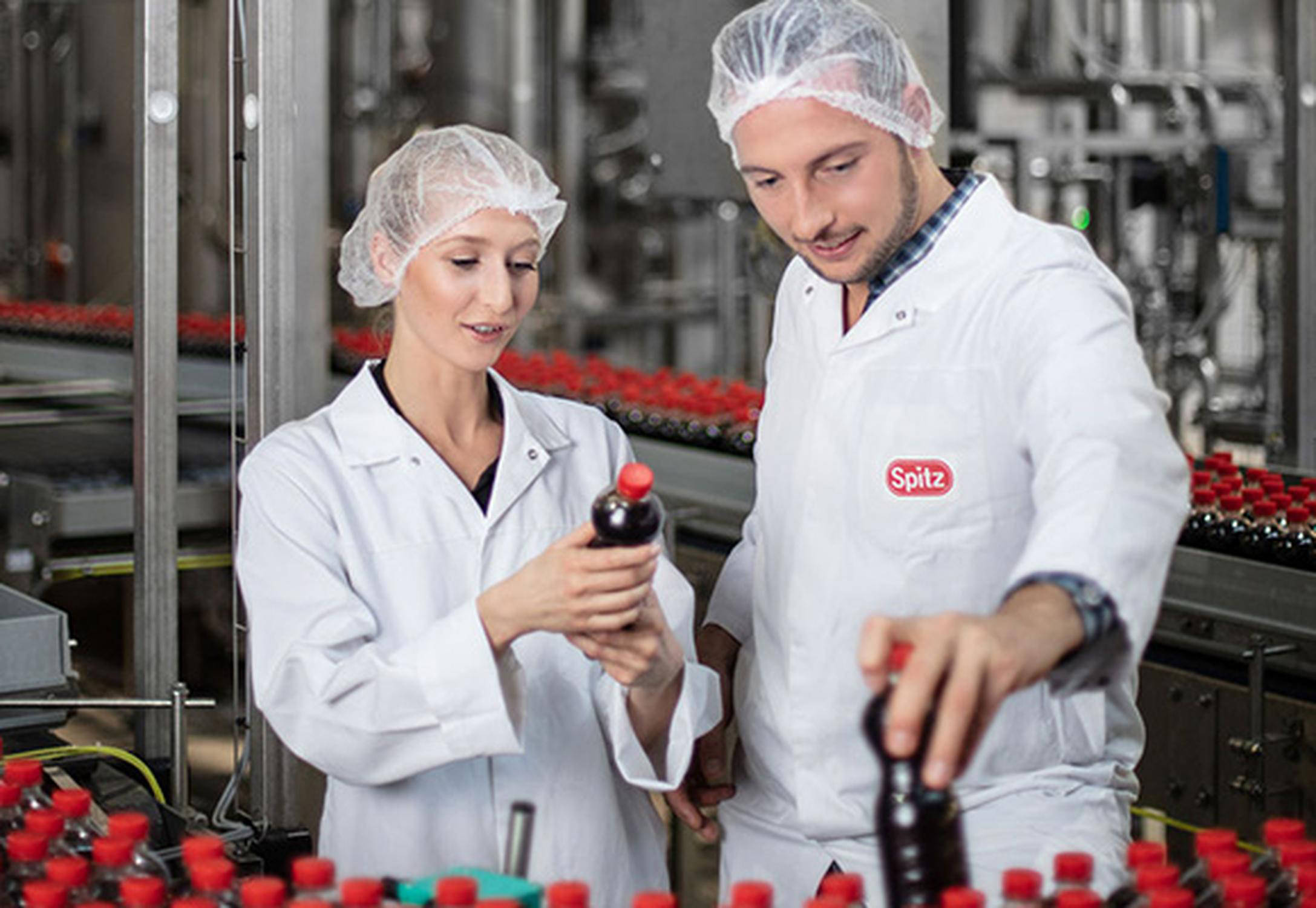 Frau und Mann in weißer Arbeitskleidung und weißem Haarnetz mit je einer Flasche in der Hand.
