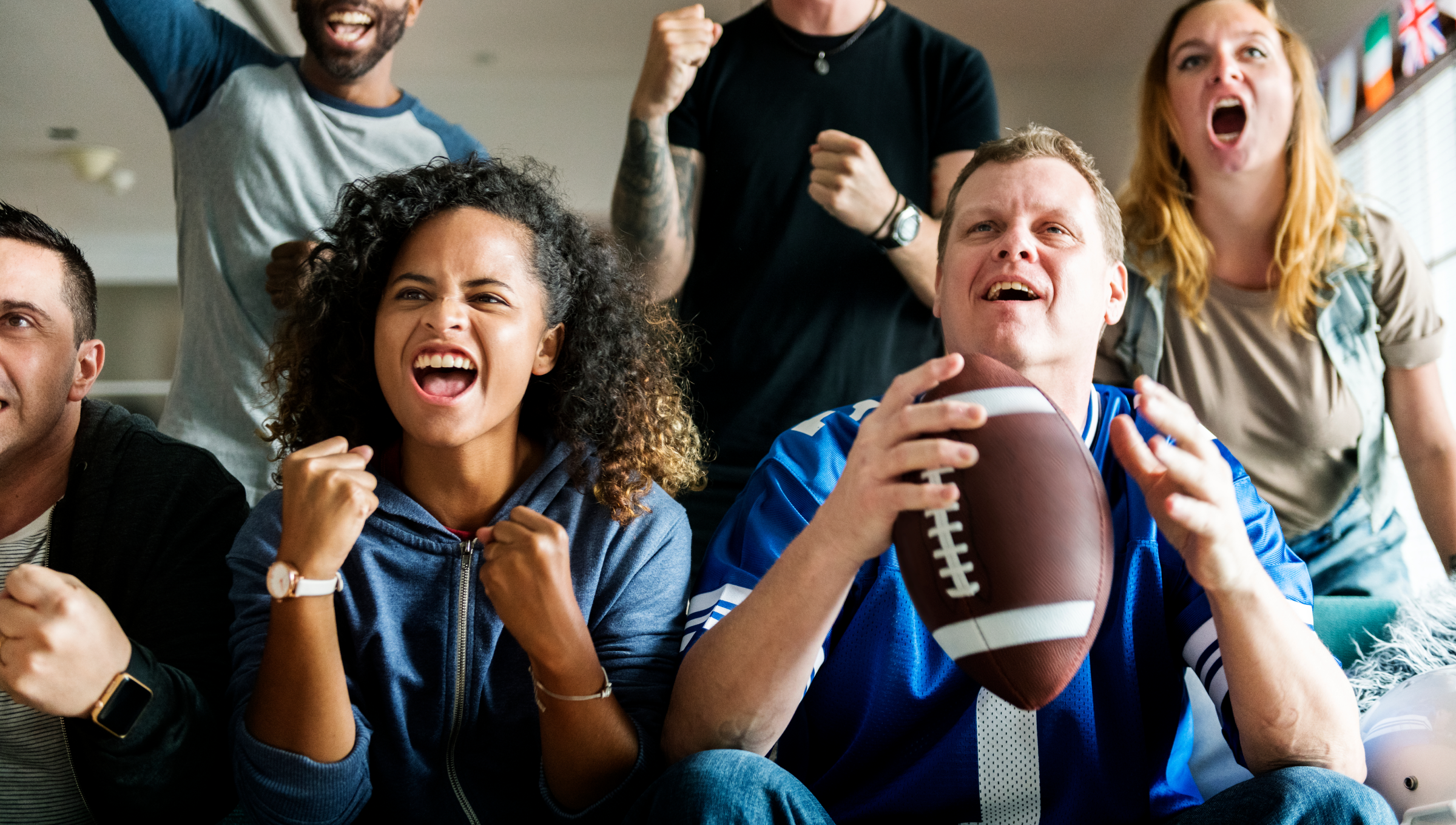Freunde schauen gemeinsam den Super Bowl und jubeln, ein Mann hält einen Football in der Hand.