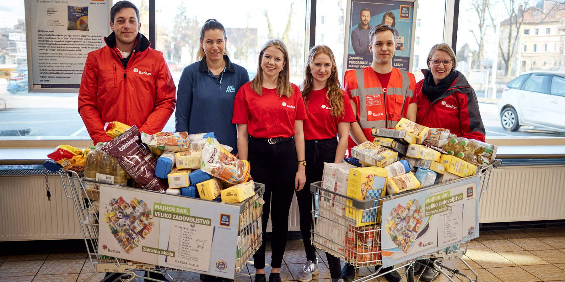 Hofer zapoleni z nakupovalnimi vozički polnimi darovane hrane, napis: Podpiramo več kot 40 nevladnih organizacij