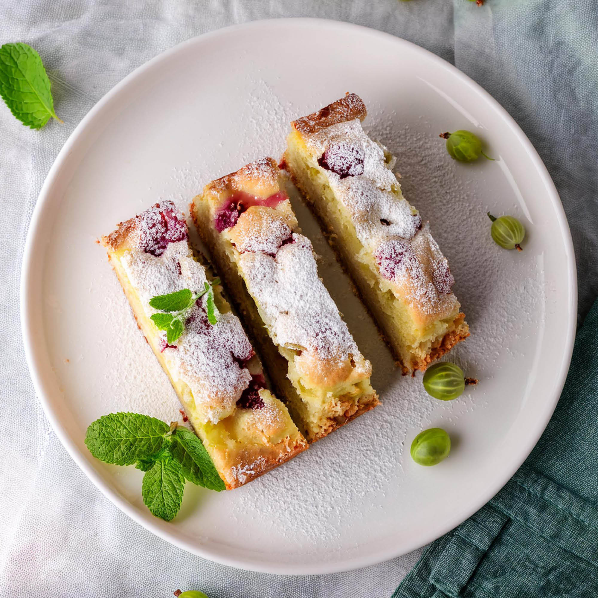 Drei Beerenkuchenstücke mit Puderzucker, Minze und Stachelbeeren.