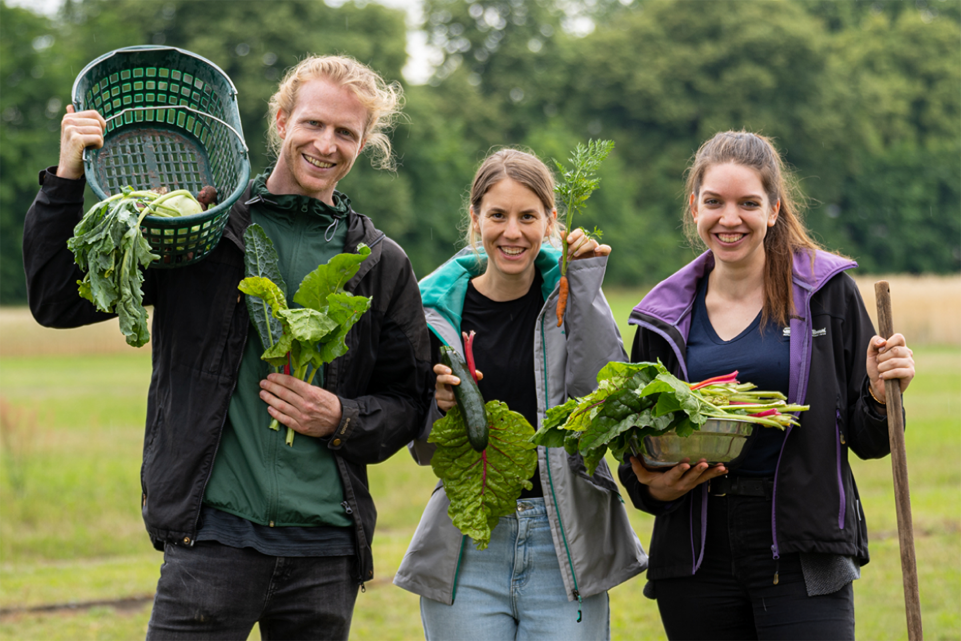 Drei Lehrkräfte halten auf einem Acker verschiedenes Gemüse in den Händen.