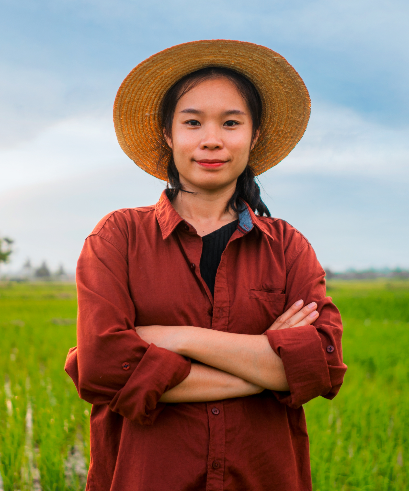 Junge Bäuerin mit Strohhut und rotem Hemd steht auf einem grünen Feld und blickt selbstbewusst in die Kamera.