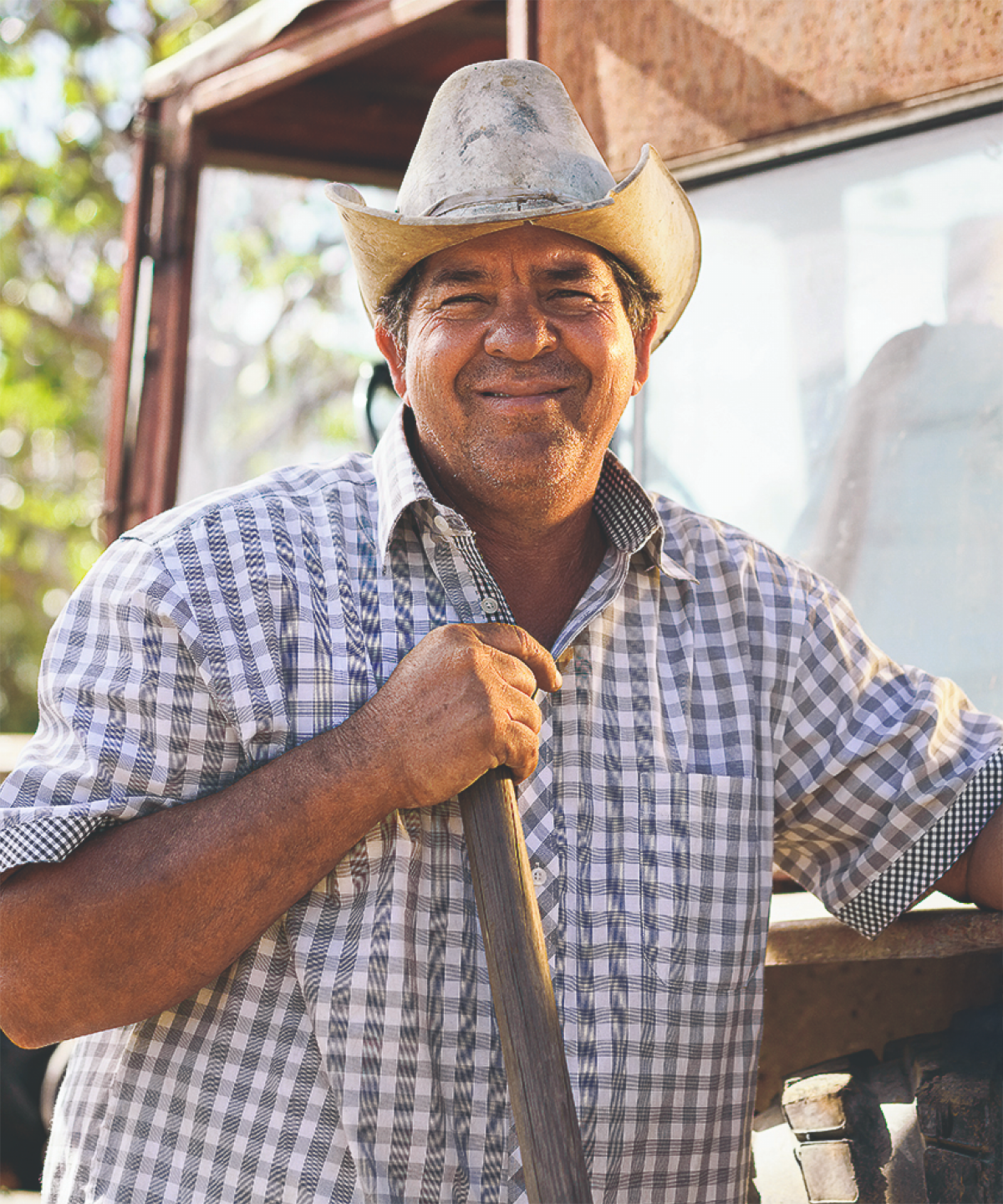 Bauer mit Hut und kariertem Hemd lächelt vor einem Traktor in einer ländlichen Umgebung.