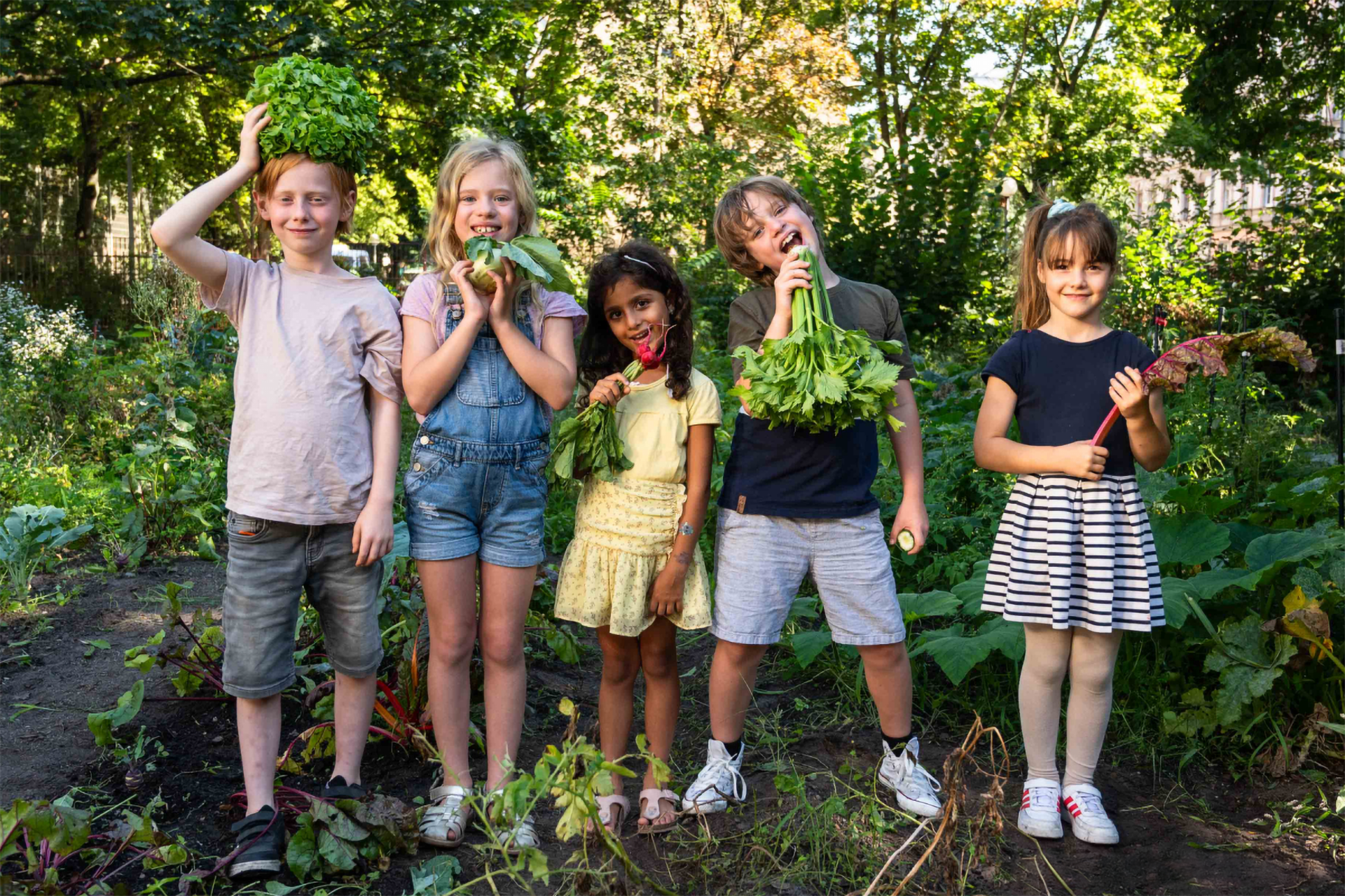 Eine Gruppe von Kindern posiert auf lustige Weise mit frisch geerntetem Gemüse auf einem Beet.