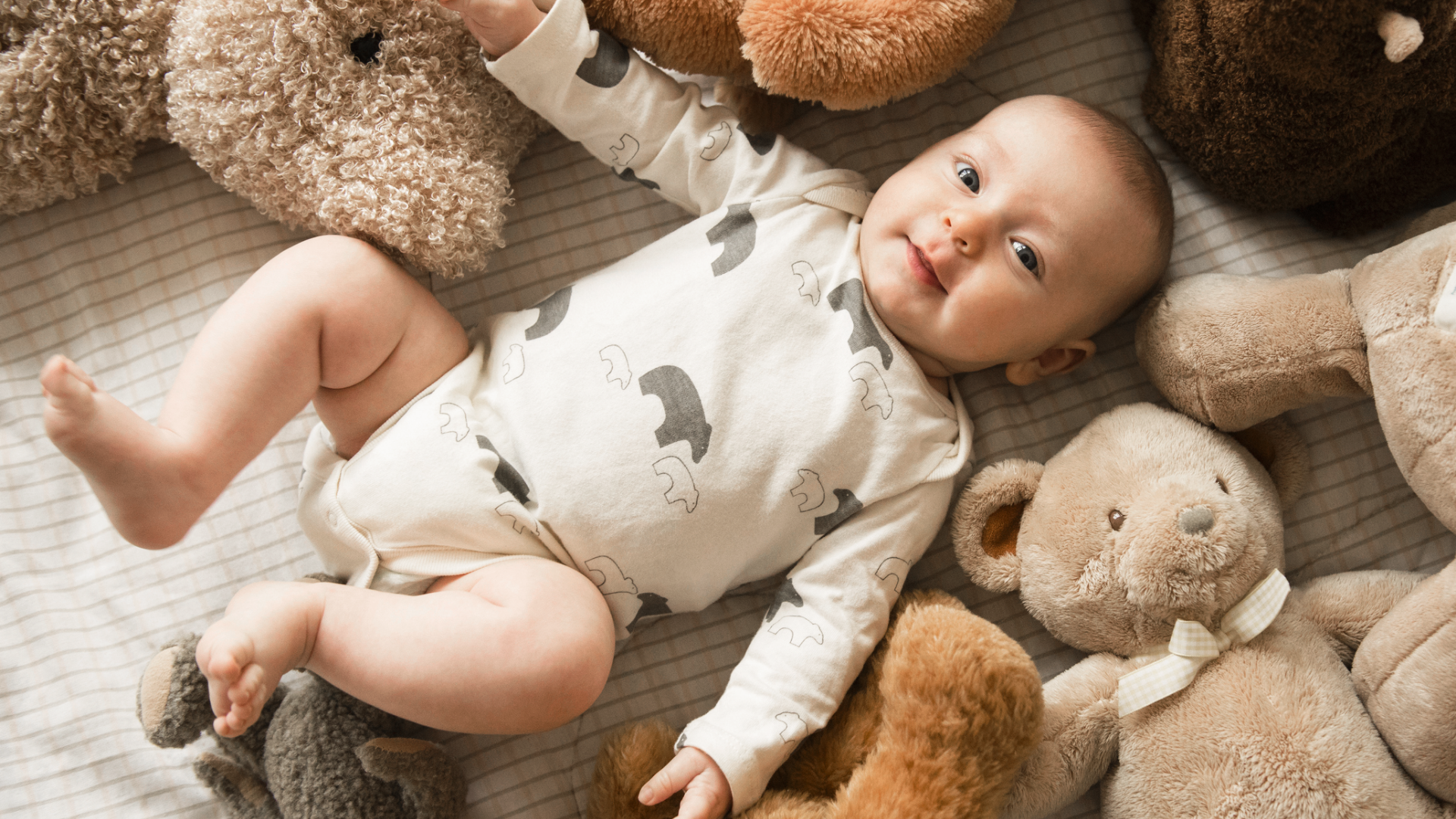Baby liegt umgeben von Teddybären auf gestreifter Decke.
