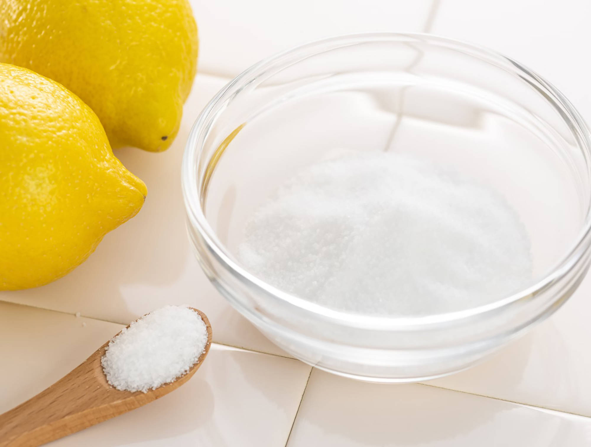 Glasschale mit Zitronensäurepulver, einem Holzlöffel und frischen Zitronen auf weißen Fliesen.