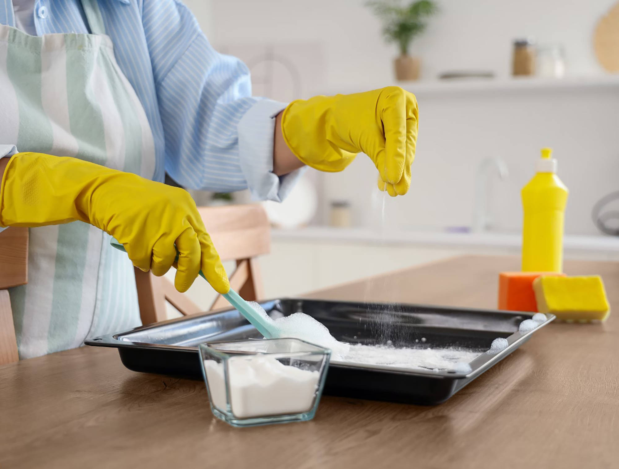 Person mit gelben Gummihandschuhen reinigt ein Backblech mit Natron und Wasser in einer modernen Küche.