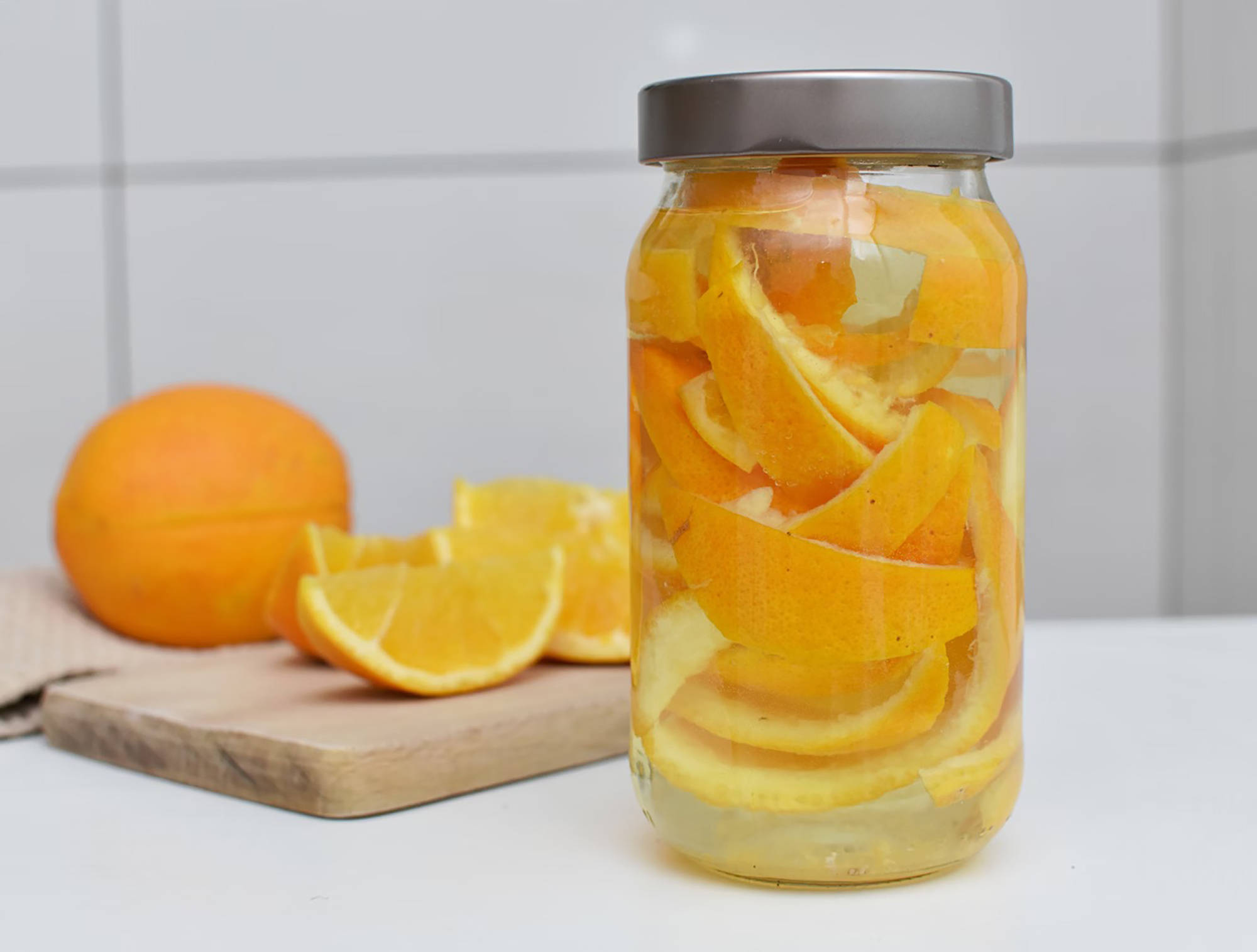 Glas mit Orangenschalen in Essig zur Herstellung eines natürlichen Reinigers, daneben frische Orangen auf einem Schneide