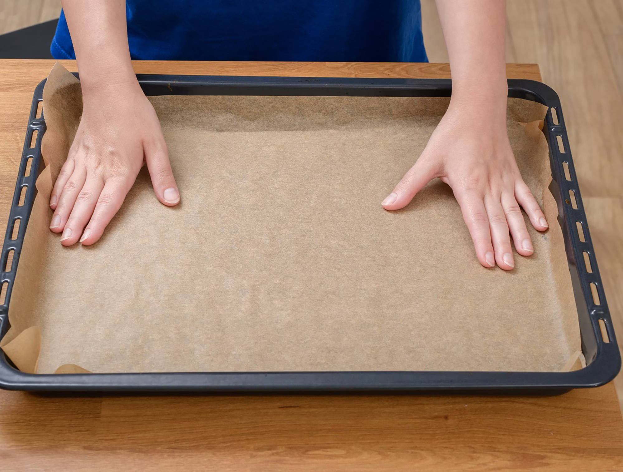 Person legt ein Stück Backpapier auf ein schwarzes Backblech, das auf einem Holztisch liegt.