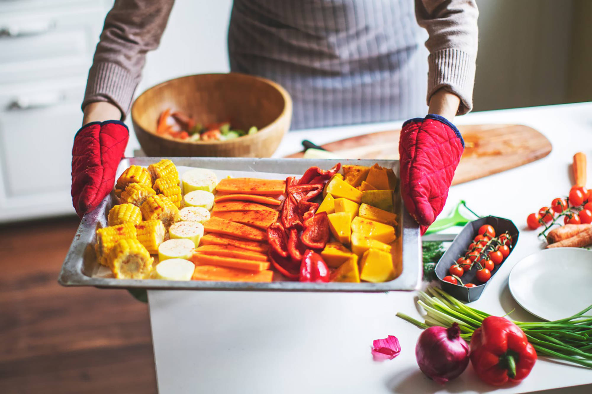 Eine Frau mit Ofenhandschuhe hält ein üppig belegtes Blech mit Ofengemüse. 