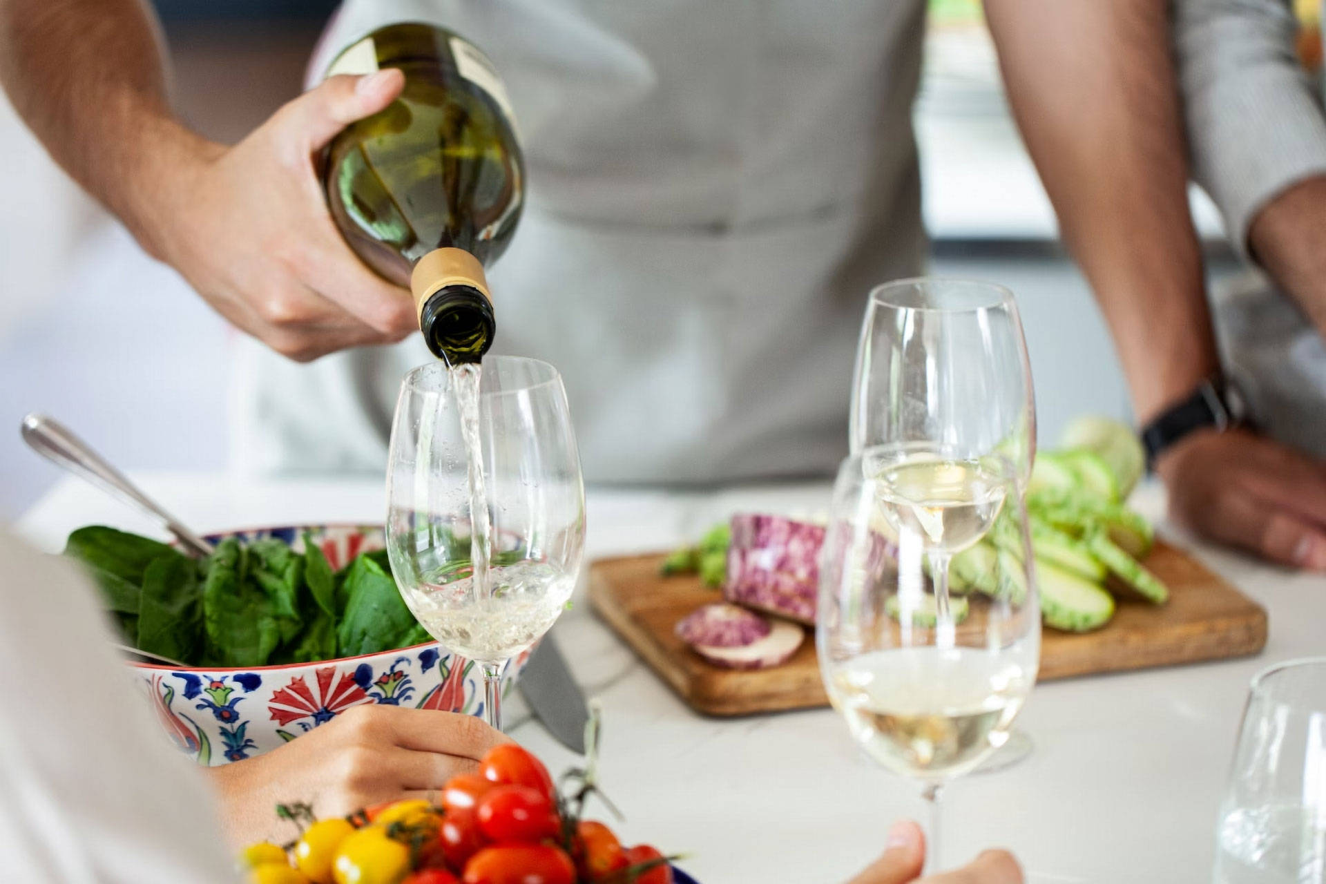 Person gießt Weißwein in ein Glas, während eine Schüssel Salat auf einem Tisch steht.