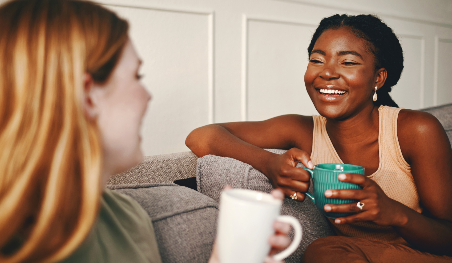 Zwei Frauen sitzen lachend mit Tassen auf dem Sofa.