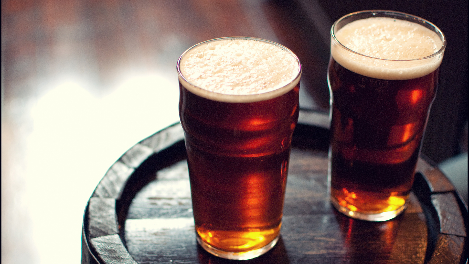 Zwei Gläser dunkles Bier mit Schaum auf Holzfass.
