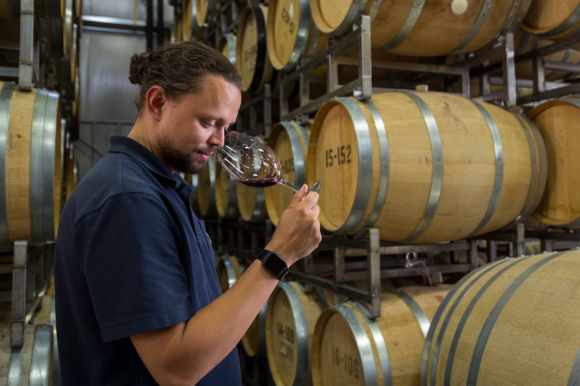 Ein Winzer prüft die Qualität des Rotweins, indem er in das Rotweinglas riecht. Hinter ihm gestapelte Weinfässer.