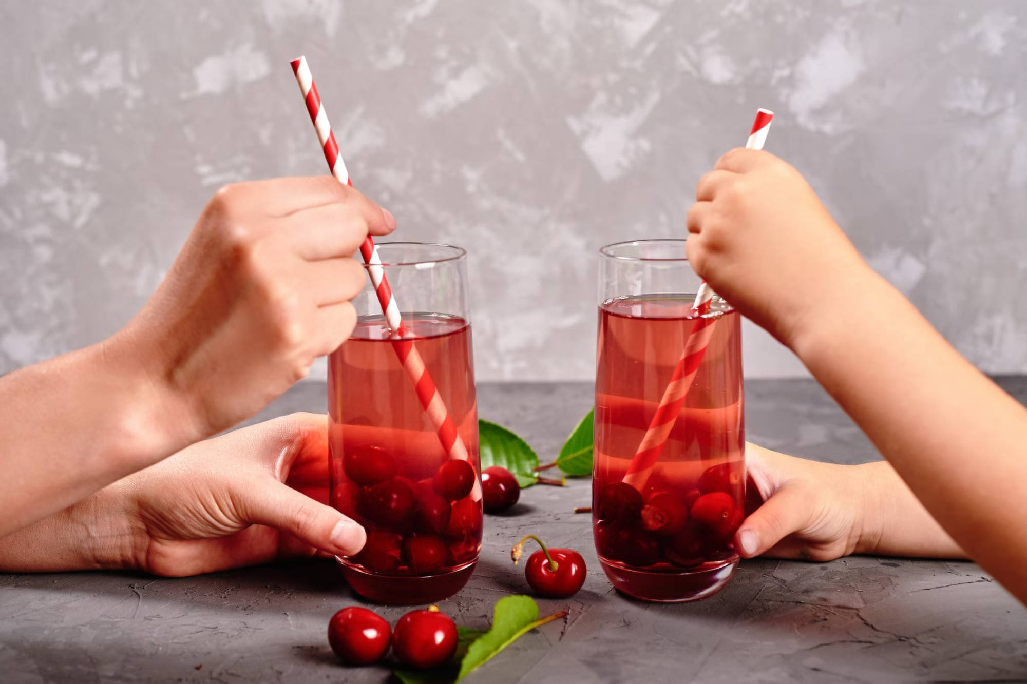 Zwei erwachsene Hände halten Krisch-Eistee mit Strohalm, gegenüber zwei Kinderhände mit gleichem Getränk. 