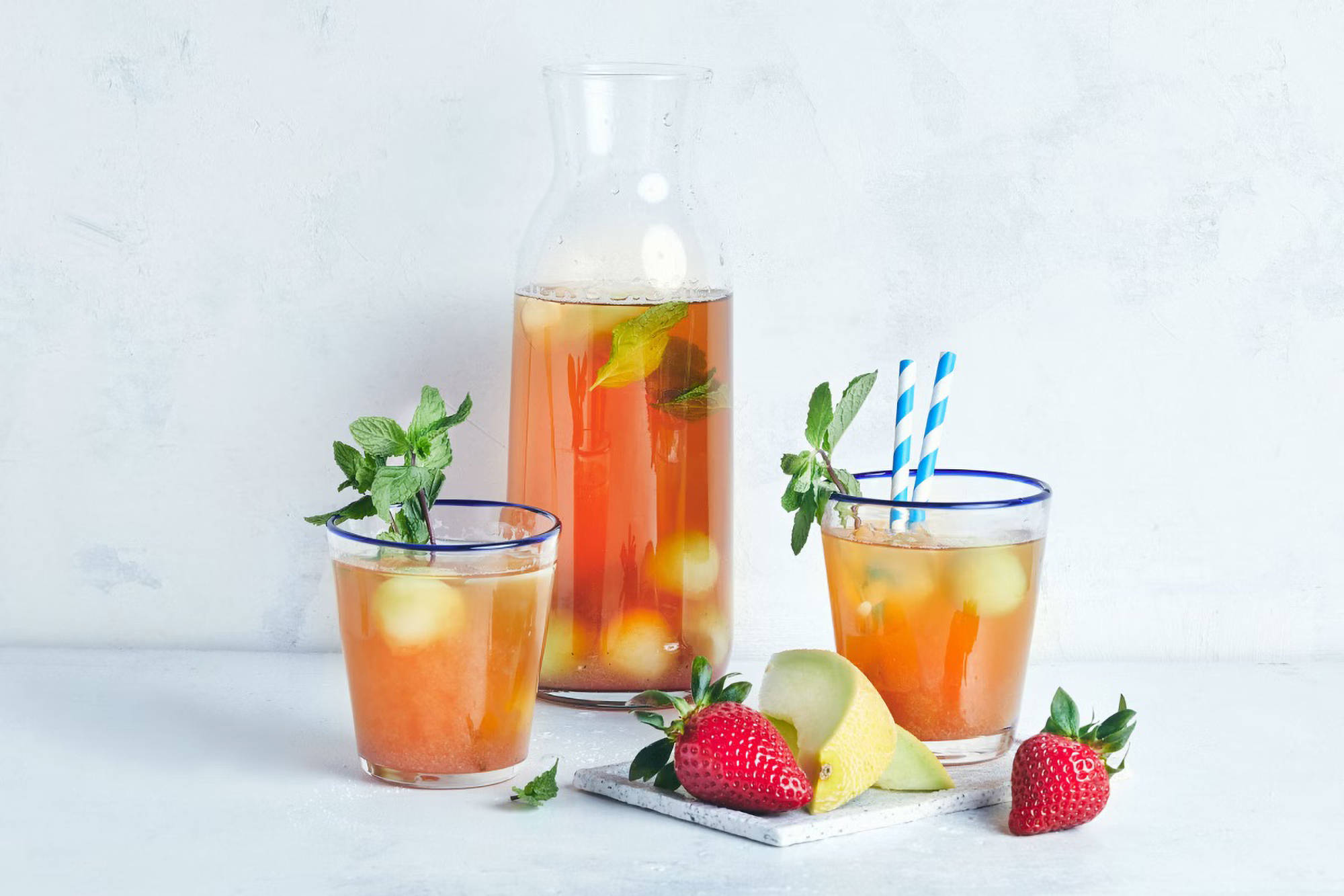 Eine Karaffe und zwei Gläser mit gelbem Eistee, Erdbeeren und Melone. 