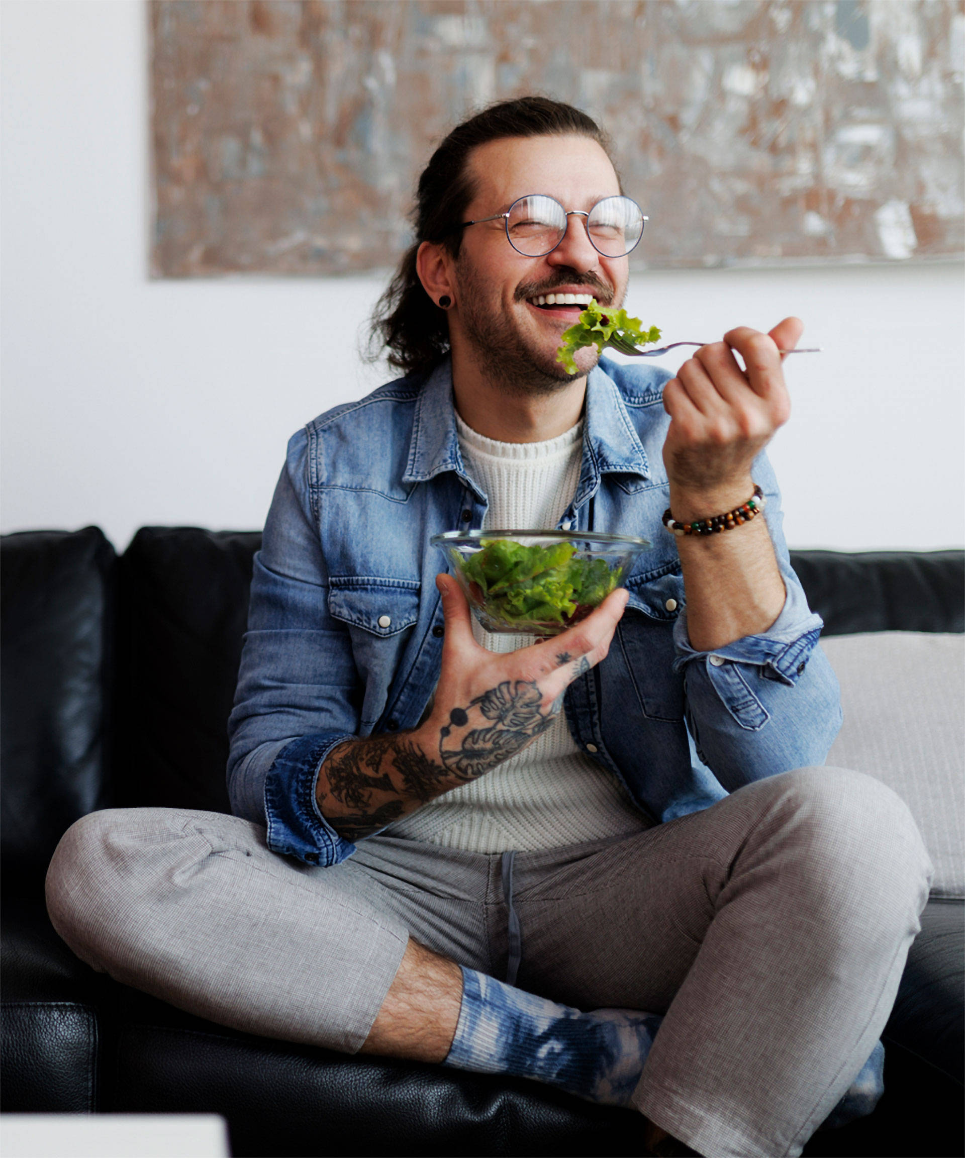 Lächelnder Mann mit Brille isst Salat aus einer Schüssel, sitzt entspannt auf einer schwarzen Couch im Wohnzimmer.