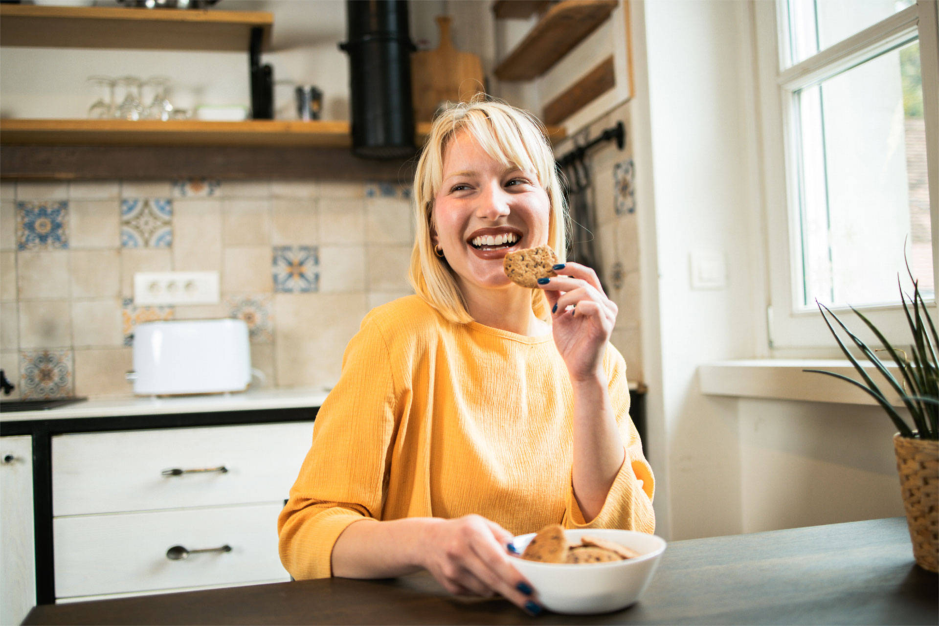 Lachende Frau in gelbem Pullover isst einen Keks aus einer Schüssel in einer gemütlichen Küche.